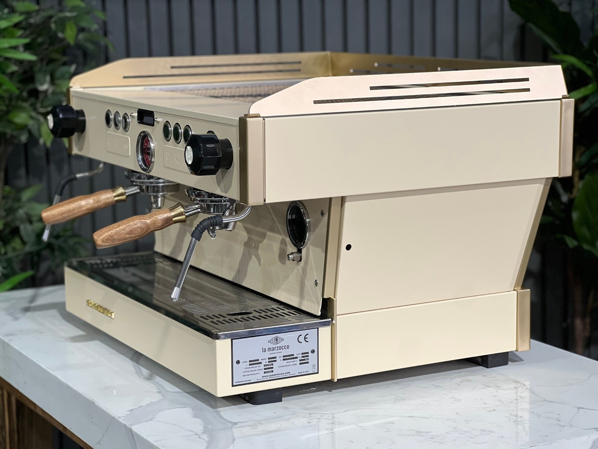 La Marzocco Linea PB 2 Group Coffee Machine Cream & Gold - Used shown at an angle on a marble bench with greenery in the background. This espresso machine features a striking cream and gold body, dual group heads, and digital controls for professional espresso performance.
