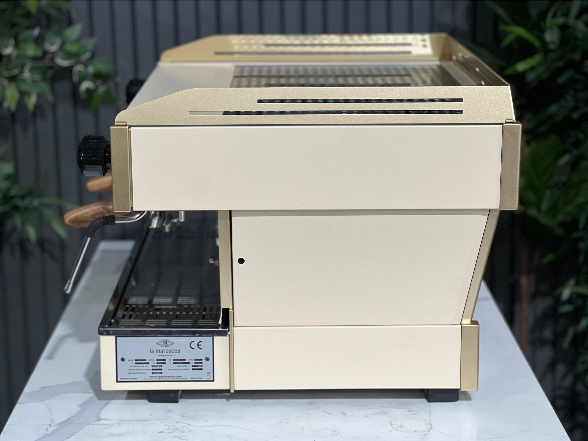 La Marzocco Linea PB 2 Group Coffee Machine Cream & Gold - Used viewed side-on on a marble bench with greenery behind. This commercial espresso machine features dual steam wands and a refined silhouette designed for high-volume espresso service.