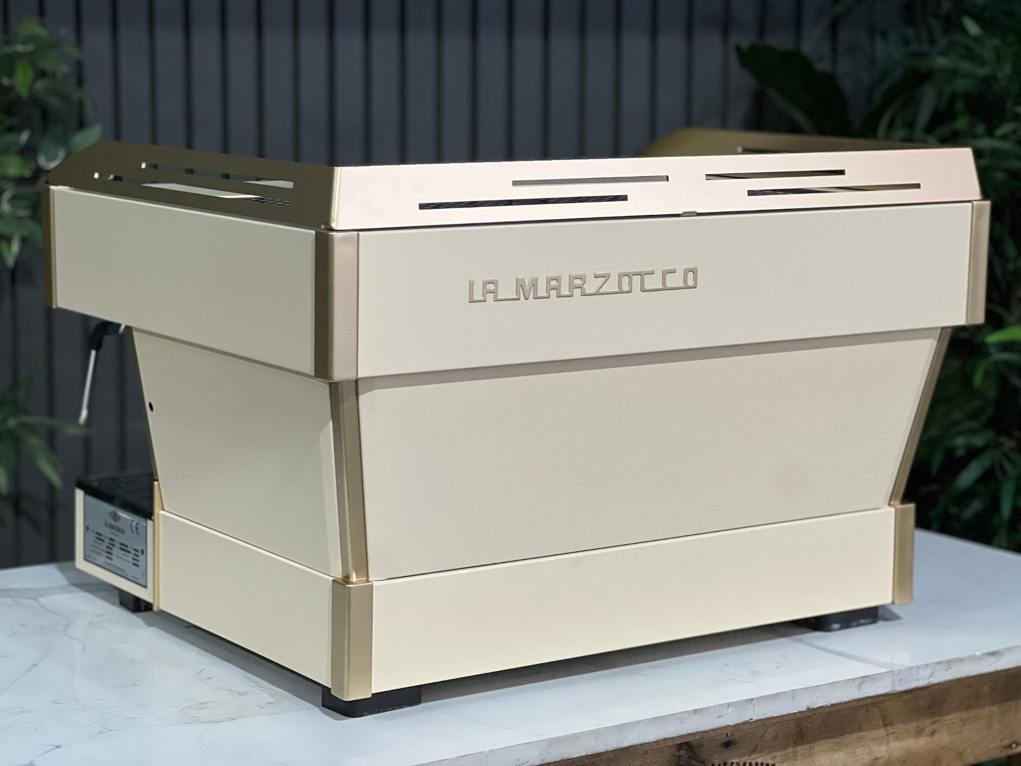 La Marzocco Linea PB 2 Group Coffee Machine Cream & Gold - Used shown at an angled back view on a marble bench with greenery. The espresso machine’s elegant design and premium build are clearly visible from this rear perspective.