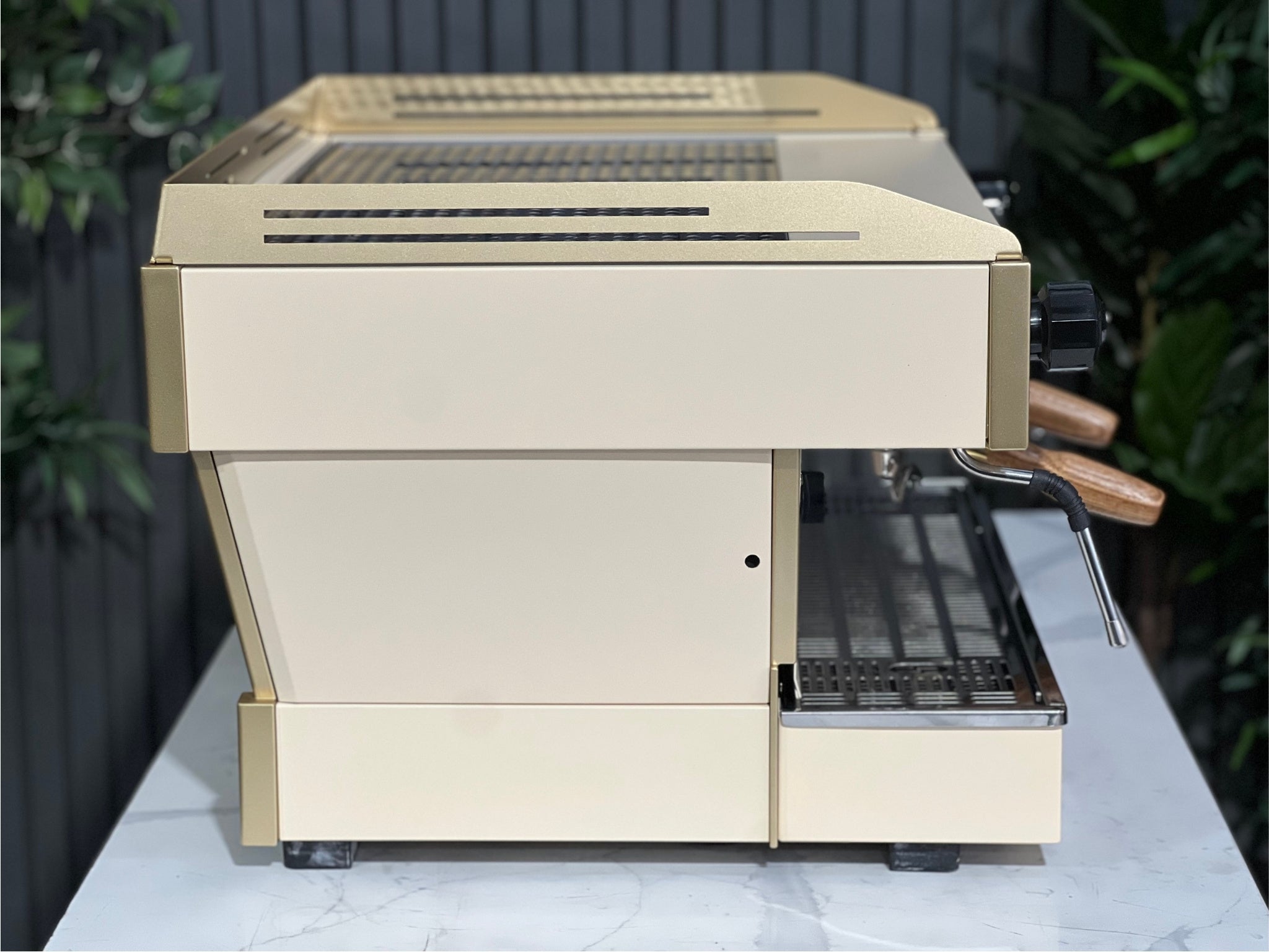 La Marzocco Linea PB 2 Group Coffee Machine Cream & Gold - Used viewed side-on on a marble bench with greenery behind. This commercial espresso machine features dual steam wands and a refined silhouette designed for high-volume espresso service.