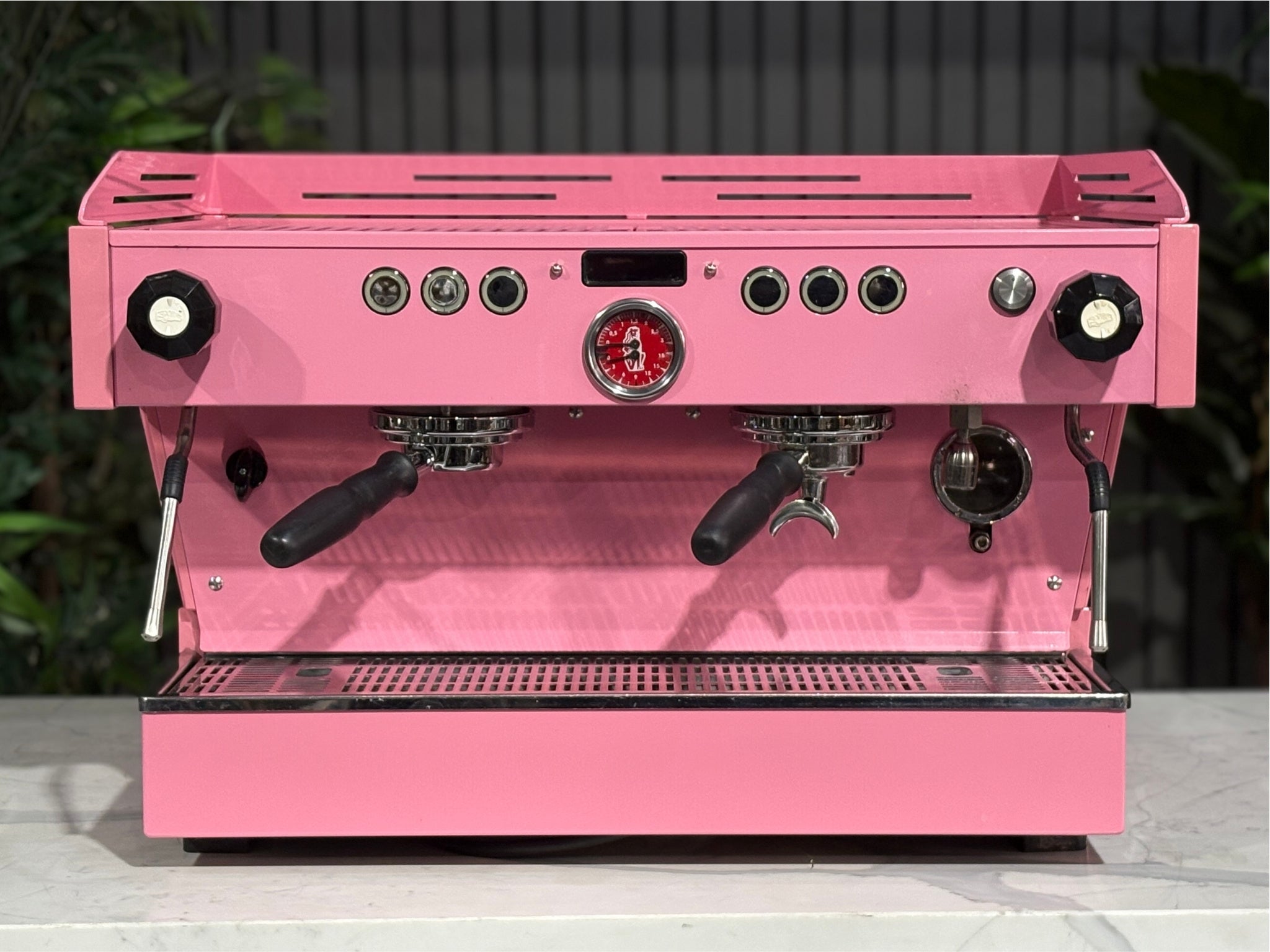 La Marzocco Linea PB 2 Group Coffee Machine Dark Pink front-on view highlighting the group heads, cup tray, and digital display, with its bold espresso-ready design standing out against a marble bench and lush green backdrop.