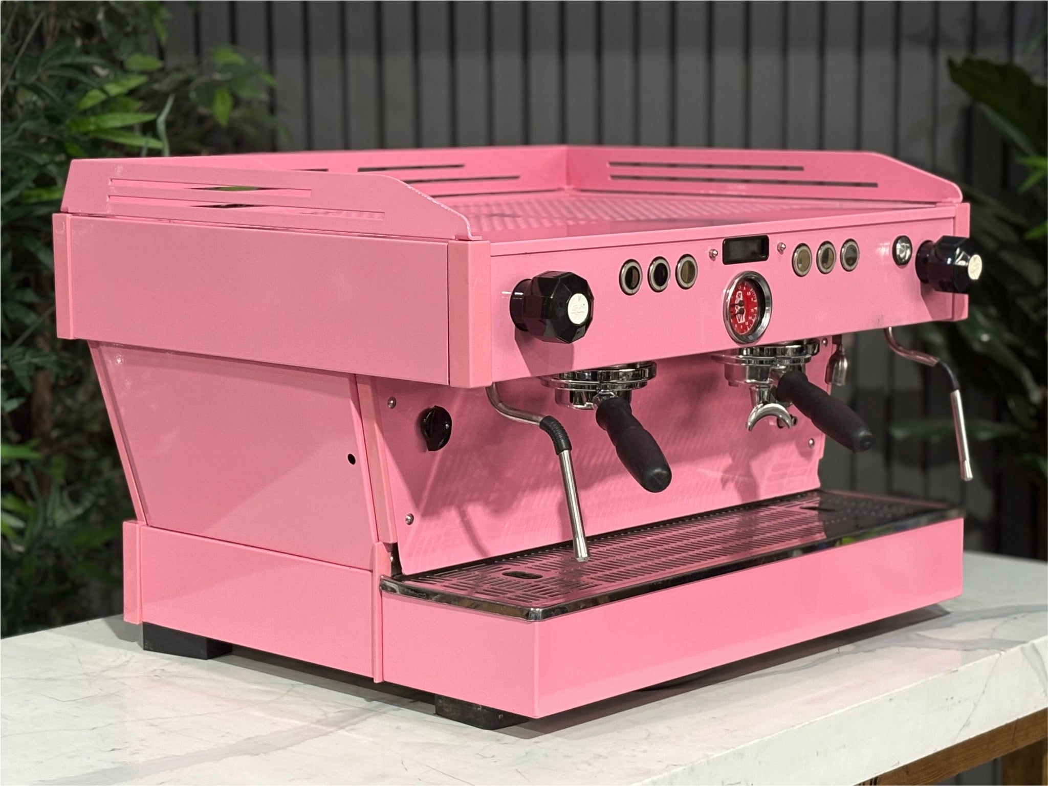 La Marzocco Linea PB 2 Group Coffee Machine Dark Pink angled view showcasing the dual group heads, LED display, and striking custom pink body of this commercial espresso coffee machine, positioned on a marble bench with greenery in the background.