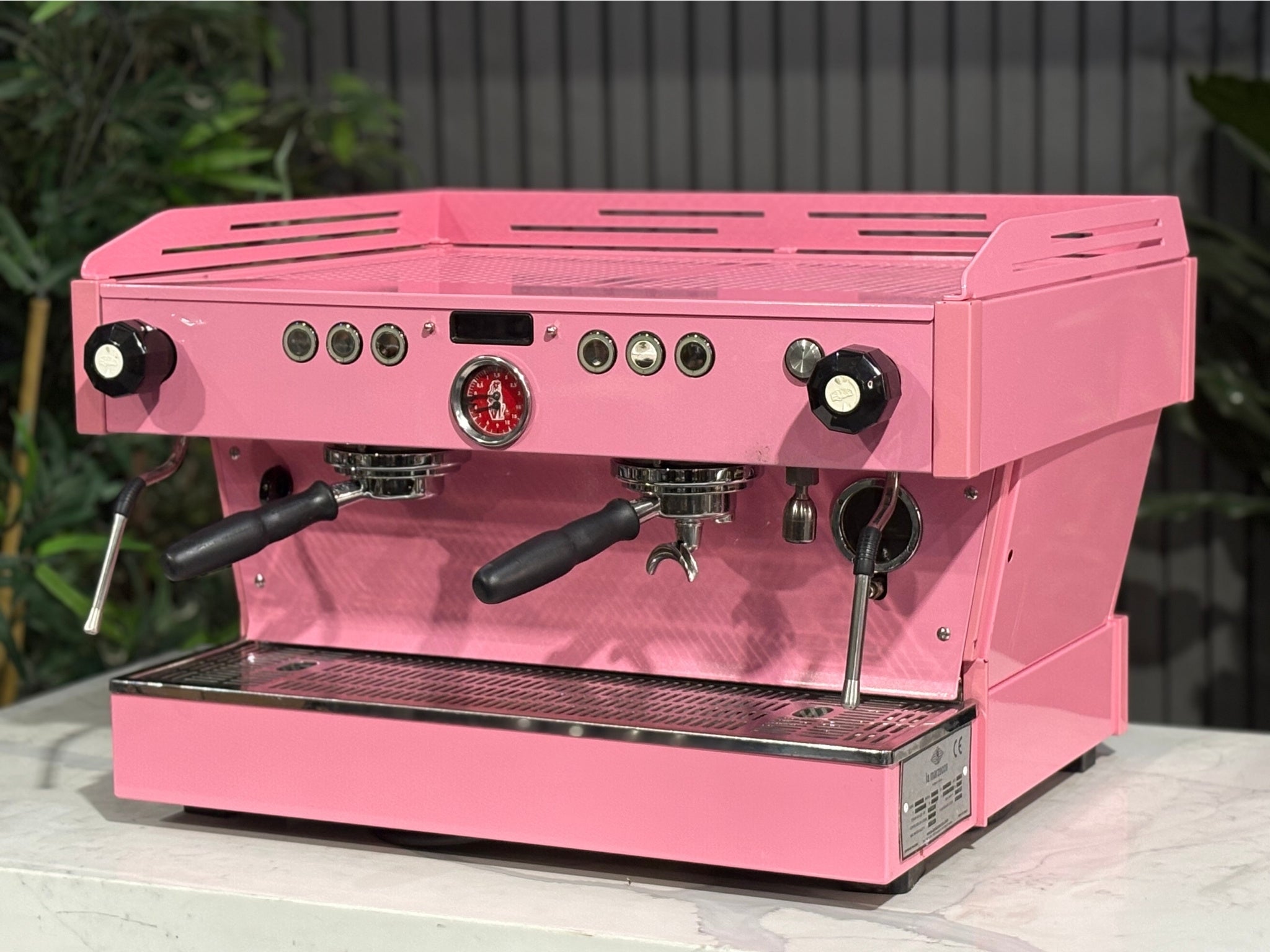 La Marzocco Linea PB 2 Group Coffee Machine Dark Pink angled view showcasing the dual group heads, LED display, and striking custom pink body of this commercial espresso coffee machine, positioned on a marble bench with greenery in the background.