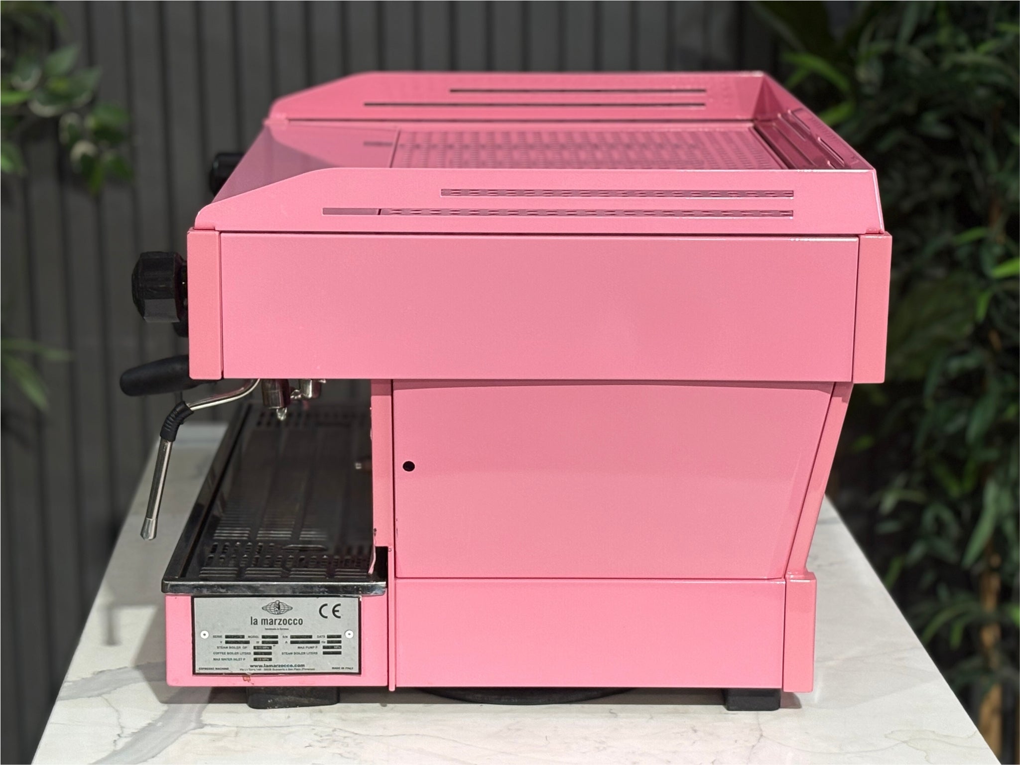 La Marzocco Linea PB 2 Group Coffee Machine Dark Pink side view revealing one group head, the dual boiler structure, and part of the steam wand, all in a vibrant pink finish on a marble bench with greenery.