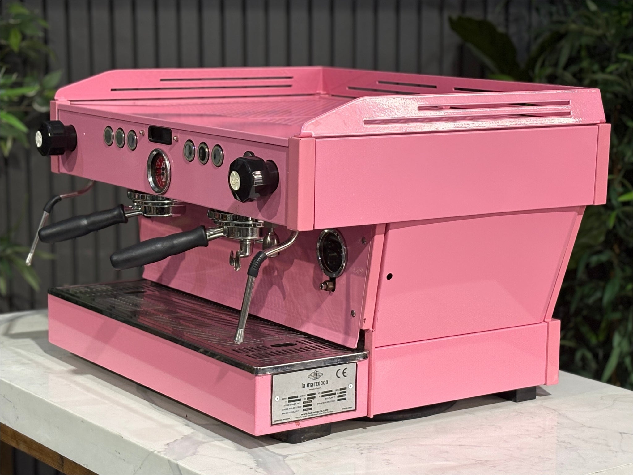 La Marzocco Linea PB 2 Group Coffee Machine Dark Pink angled view showcasing the dual group heads, LED display, and striking custom pink body of this commercial espresso coffee machine, positioned on a marble bench with greenery in the background.