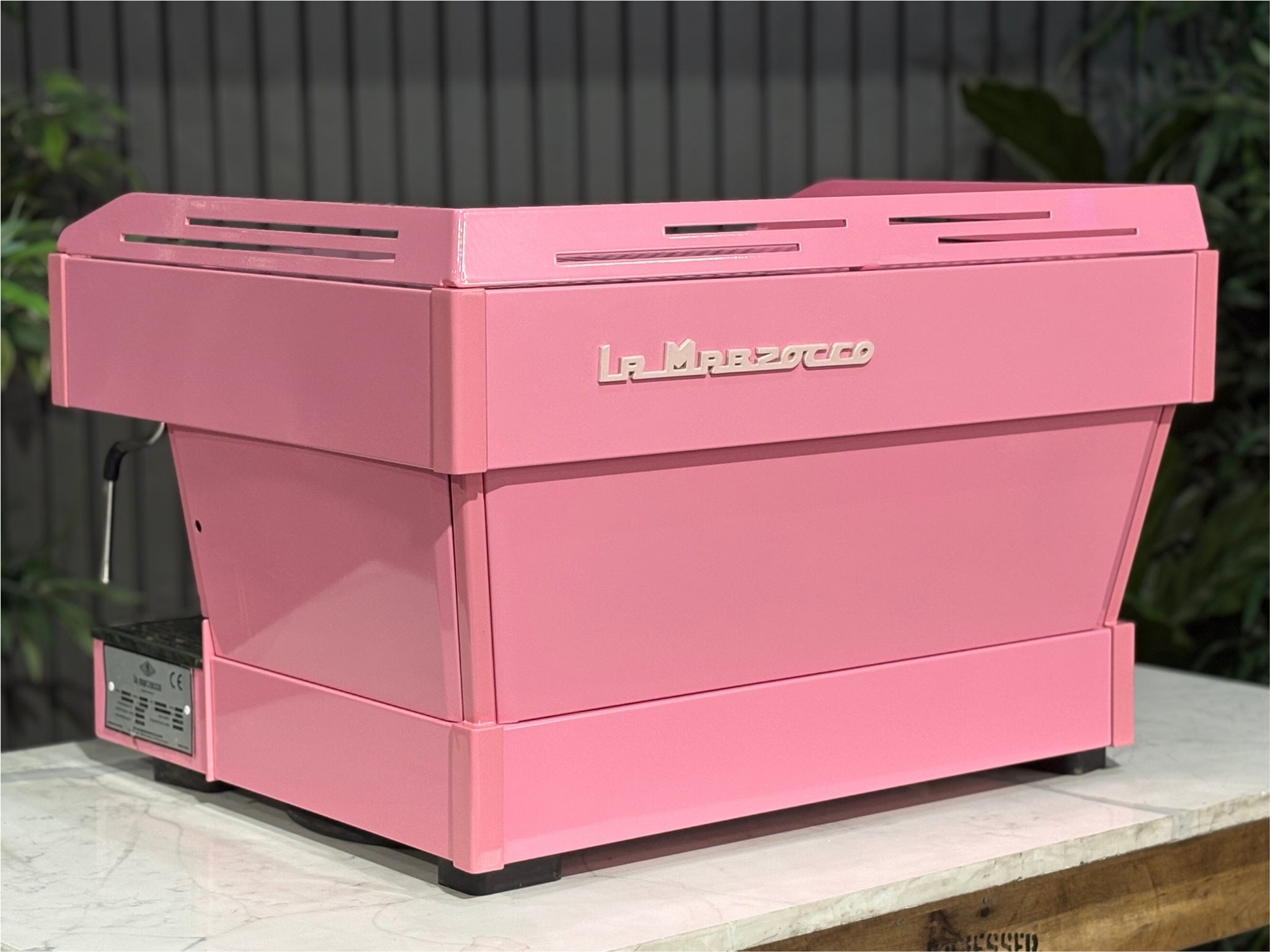 La Marzocco Linea PB 2 Group Coffee Machine Dark Pink rear-angled view showing the sleek lines of the custom pink body and rear panel, ideal for espresso performance in a café setting, with greenery behind.