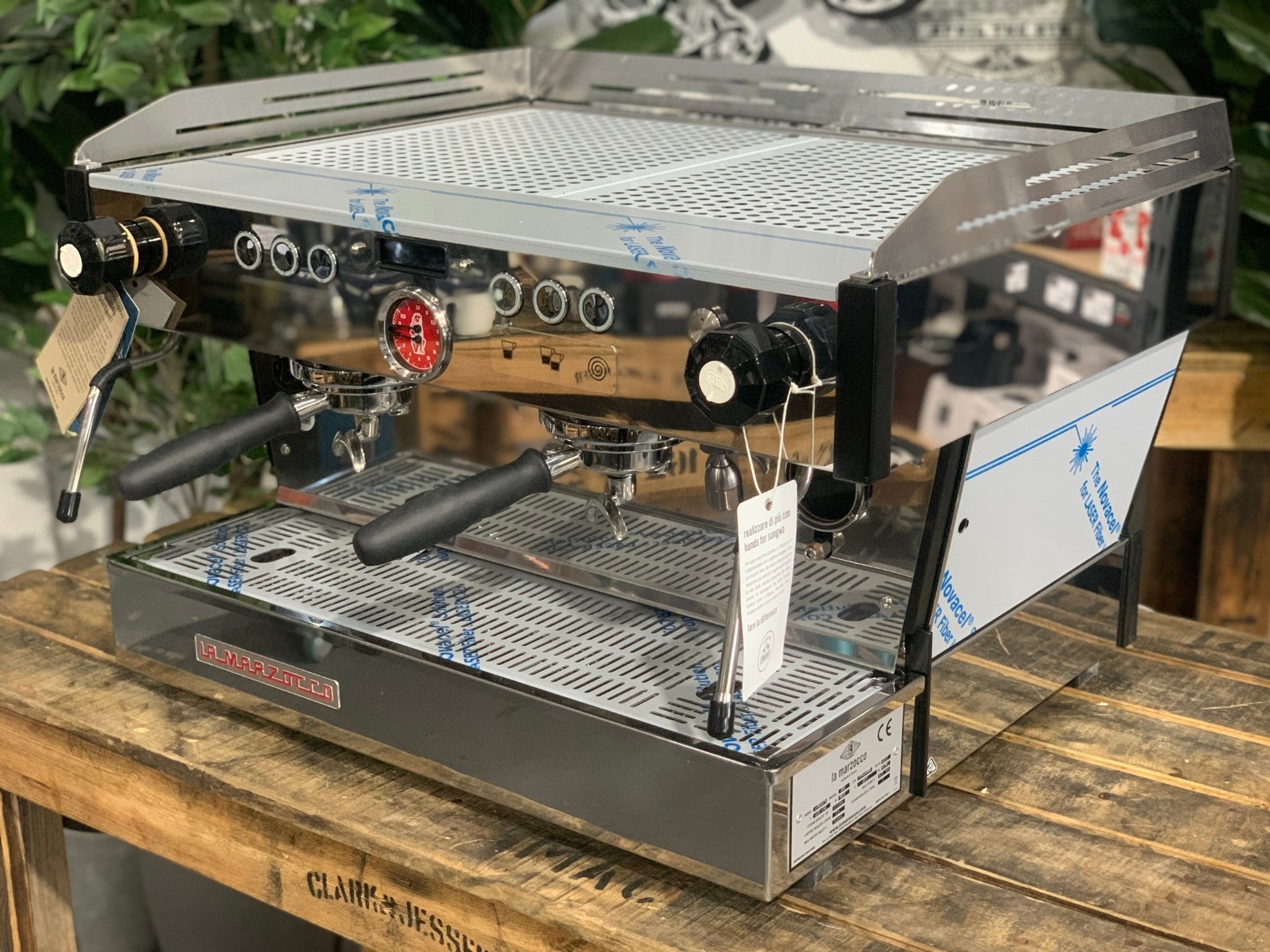 La Marzocco Linea PB 2 Group Coffee Machine Stainless – New shown at an angled view on a wooden bench with greenery in the background, highlighting its stainless steel body, dual group heads, dual steam wands, and digital LCD display for professional espresso features.