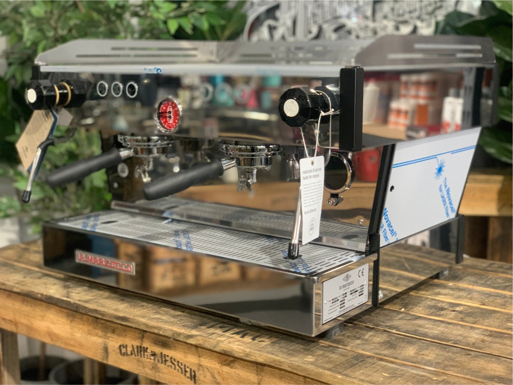 La Marzocco Linea PB 2 Group Coffee Machine Stainless – New shown at an angled view on a wooden bench with greenery in the background, highlighting its stainless steel body, dual group heads, dual steam wands, and digital LCD display for professional espresso features.