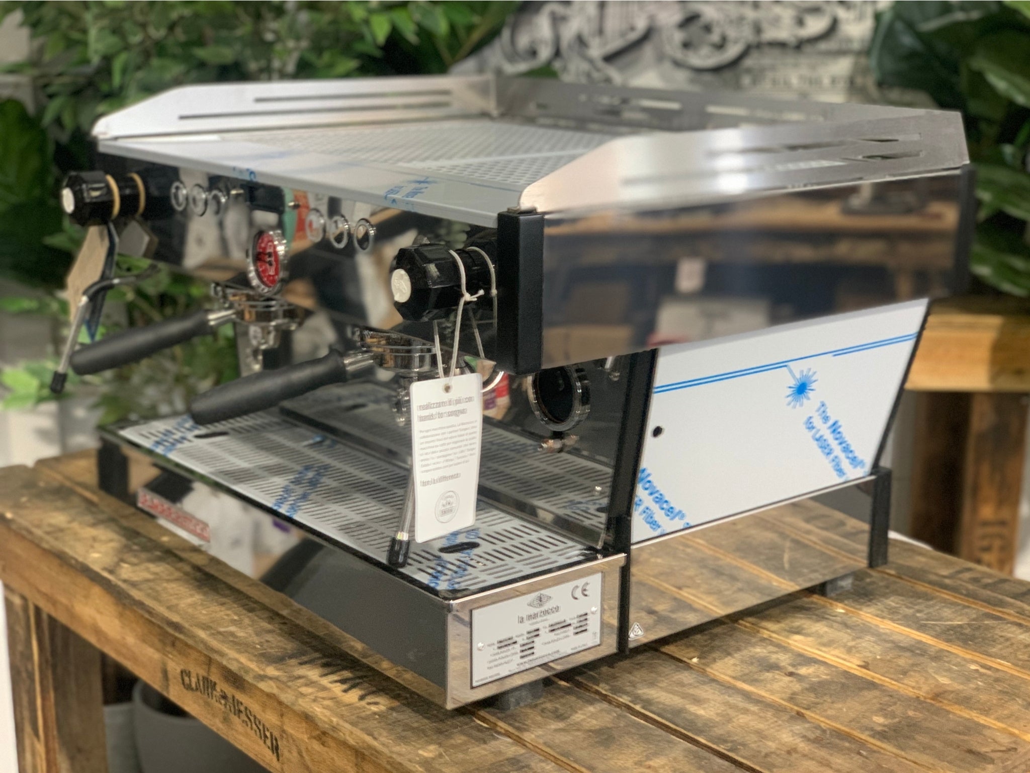 La Marzocco Linea PB 2 Group Coffee Machine Stainless – New shown at an angled view on a wooden bench with greenery in the background, highlighting its stainless steel body, dual group heads, dual steam wands, and digital LCD display for professional espresso features.