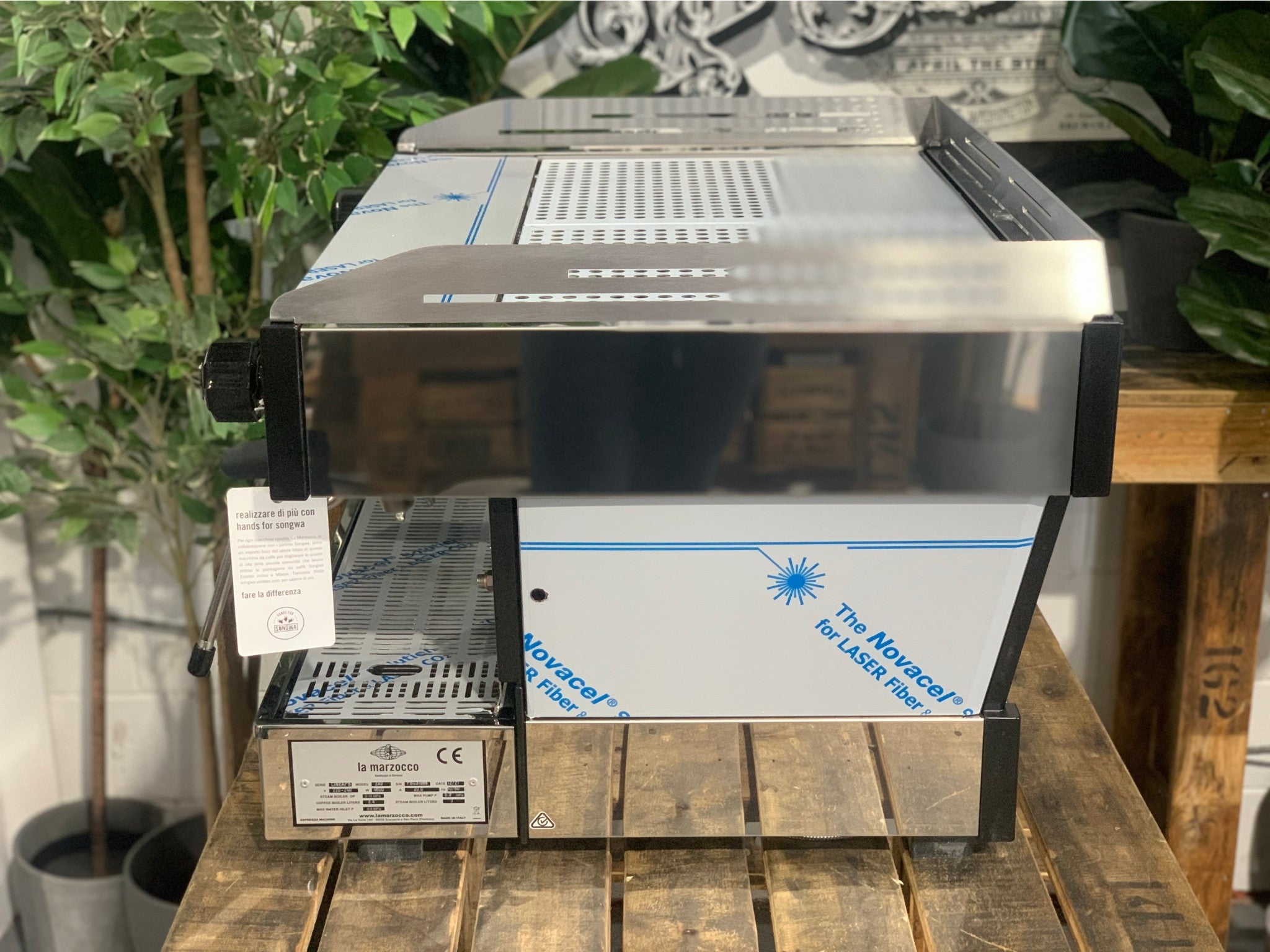 La Marzocco Linea PB 2 Group Coffee Machine Stainless – New side view on a wooden bench with greenery in the background, highlighting the stainless steel body and dual steam wands for professional espresso use.