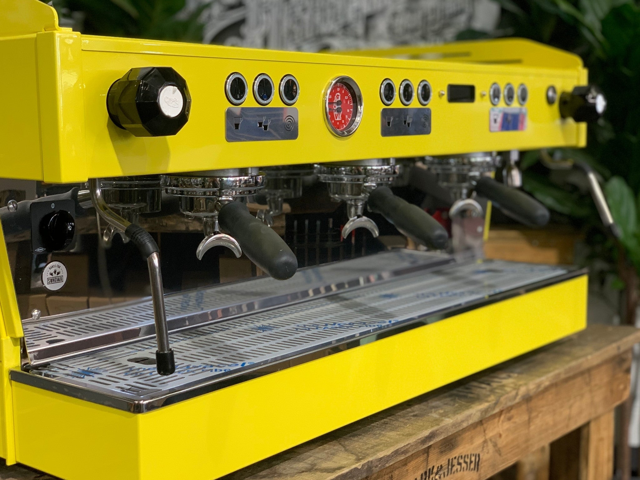 La Marzocco Linea PB 3 Group Coffee Machine Yellow – New espresso coffee machine close-up angled view on wooden bench with greenery, focusing on group heads, steam wand, cup tray, and yellow body.