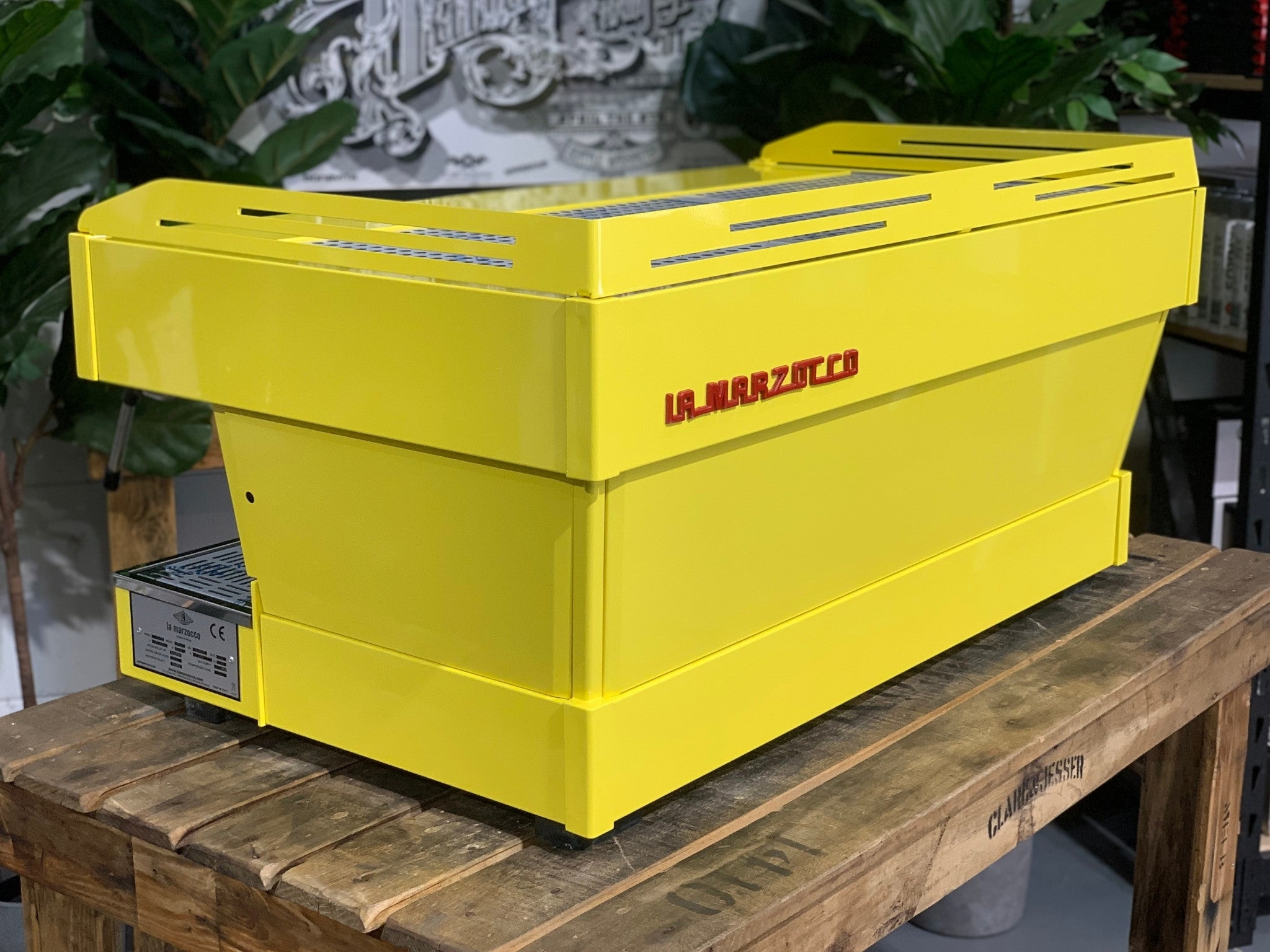 La Marzocco Linea PB 3 Group Coffee Machine Yellow – New espresso coffee machine angled rear view on wooden bench with greenery, showing yellow body, stainless trim, and machine profile.