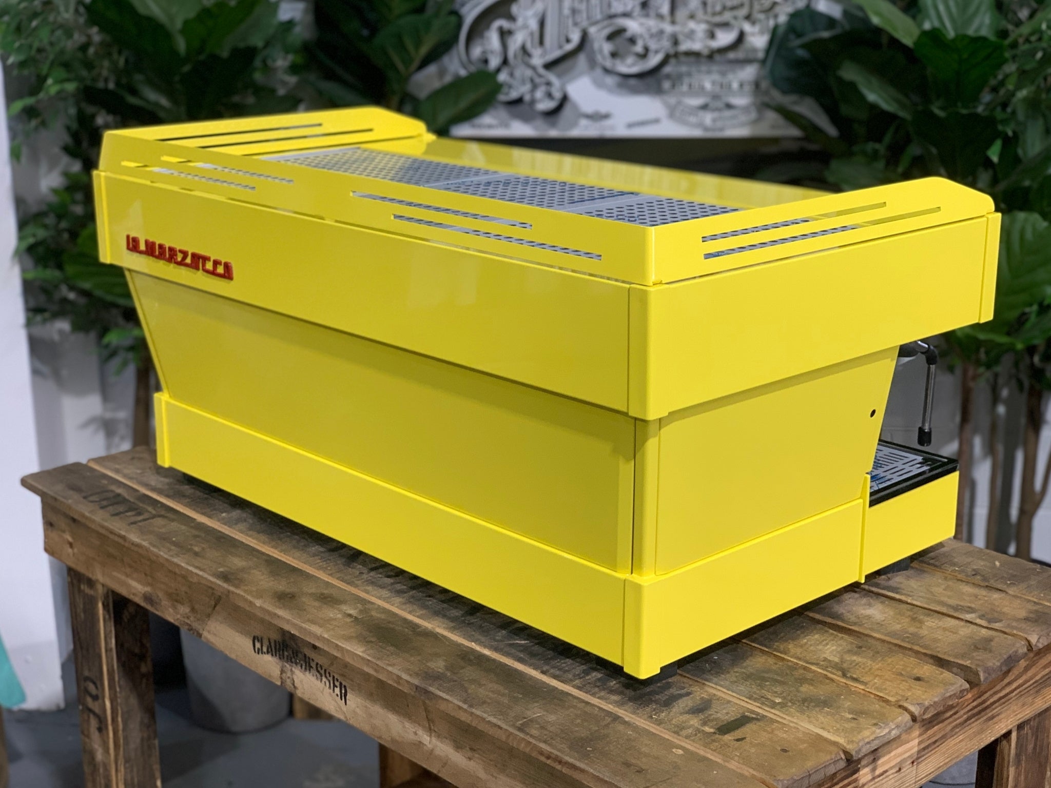 La Marzocco Linea PB 3 Group Coffee Machine Yellow – New espresso coffee machine angled rear view on wooden bench with greenery, highlighting yellow body and red La Marzocco logo.