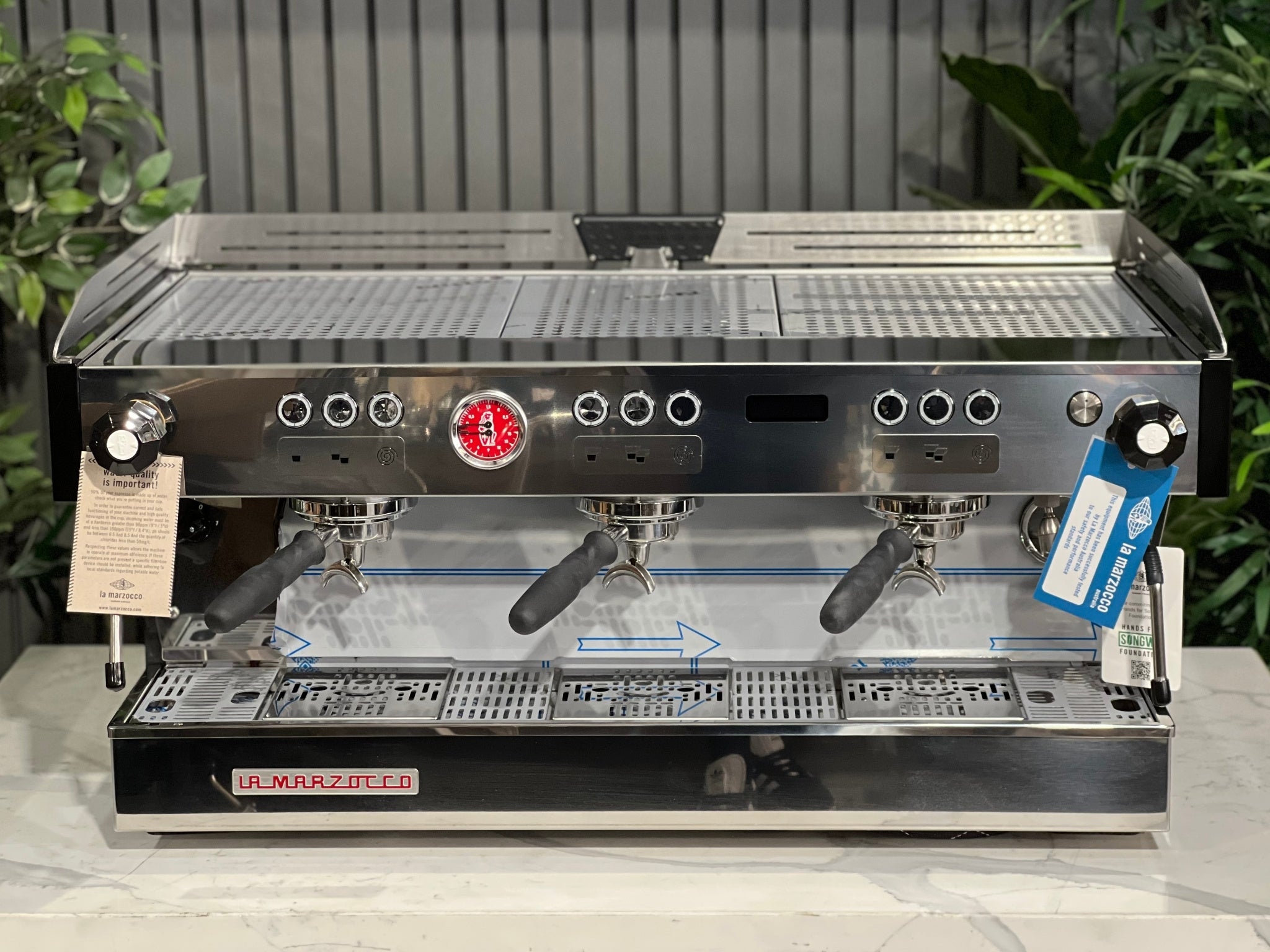 La Marzocco Linea PB ABR 3 Group Coffee Machine Stainless - New, front-on top view showing stainless steel body, cup tray, digital display, shot timers, and barista lights, on a marble bench with greenery behind.