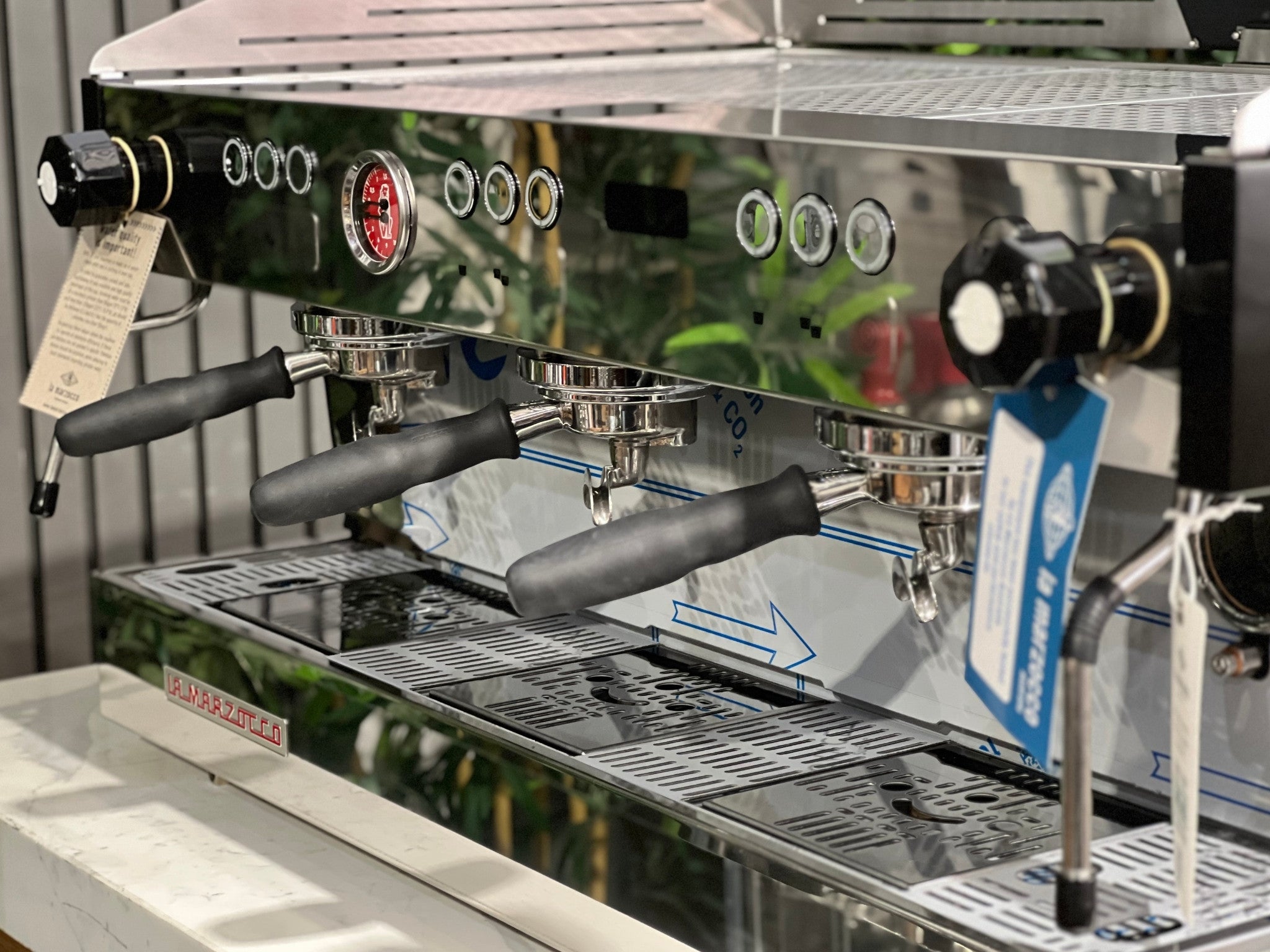 La Marzocco Linea PB ABR 3 Group Coffee Machine Stainless - New, close-up angled view of three group heads, ABR drip tray, and dual steam wands, highlighting stainless steel finish on a marble bench with greenery in the background.