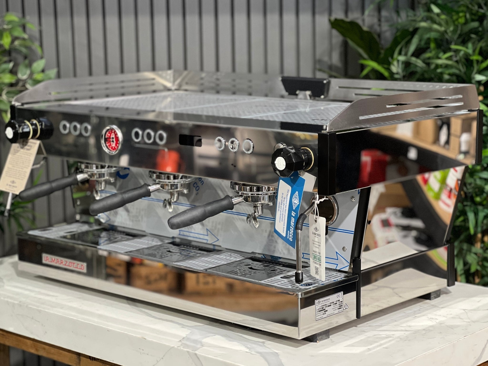 La Marzocco Linea PB ABR 3 Group Coffee Machine Stainless - New, angled view showing stainless steel body, three group heads, dual steam wands, integrated ABR drip tray, digital display, and barista lights, on a marble bench with greenery in the background.