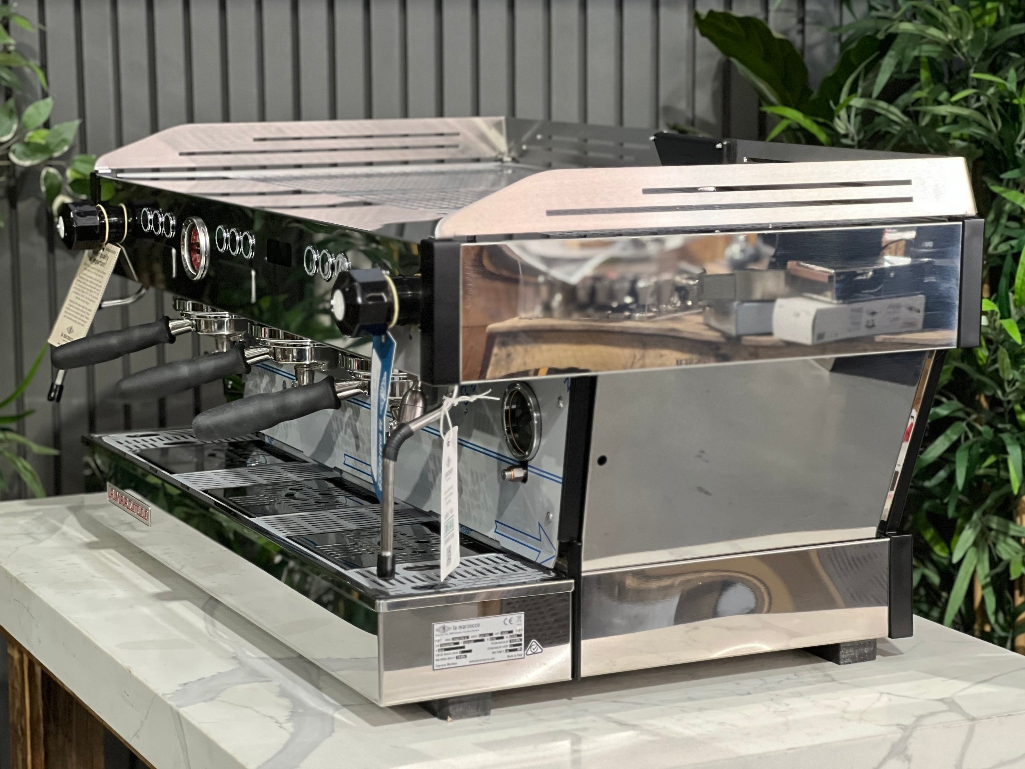 La Marzocco Linea PB ABR 3 Group Coffee Machine Stainless - New, angled view showing stainless steel body, three group heads, dual steam wands, integrated ABR drip tray, digital display, and barista lights, on a marble bench with greenery in the background.