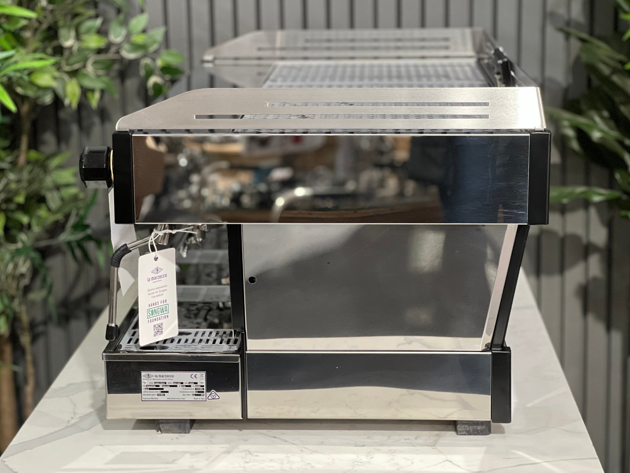 La Marzocco Linea PB ABR 3 Group Coffee Machine Stainless - New, side view showing stainless steel body, dual steam wands, and integrated ABR drip tray, set on a marble bench with greenery behind.