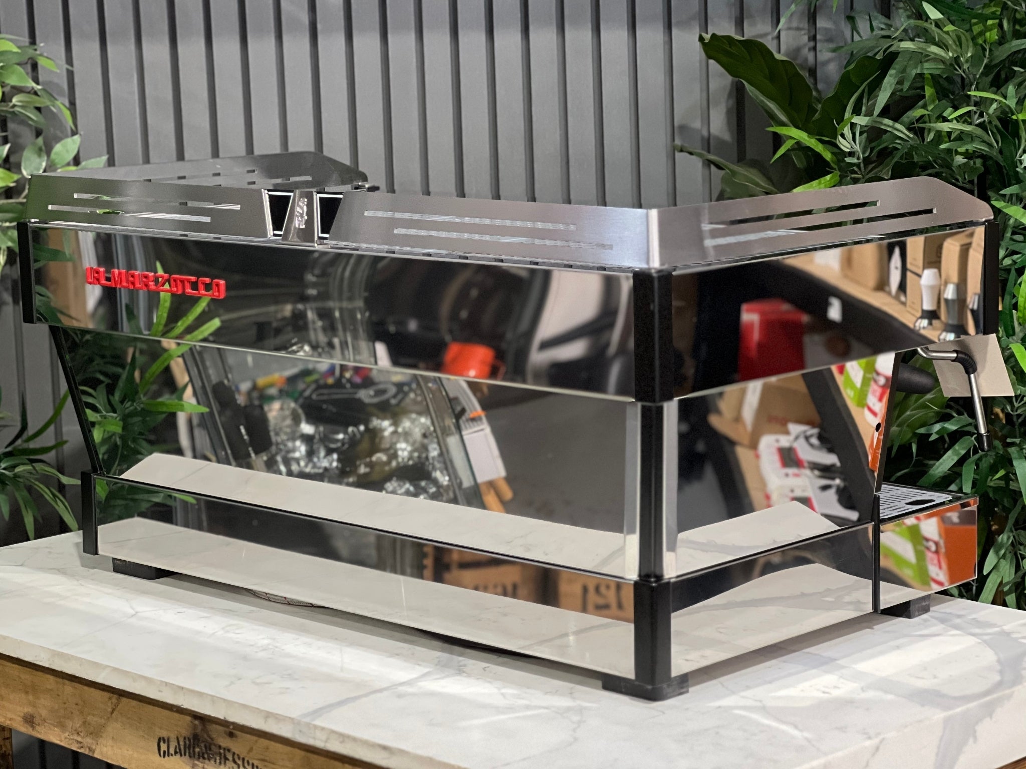 La Marzocco Linea PB ABR 3 Group Coffee Machine Stainless - New, angled back view highlighting stainless steel body, red La Marzocco logo, and side panel details, on a marble bench with greenery in the background.