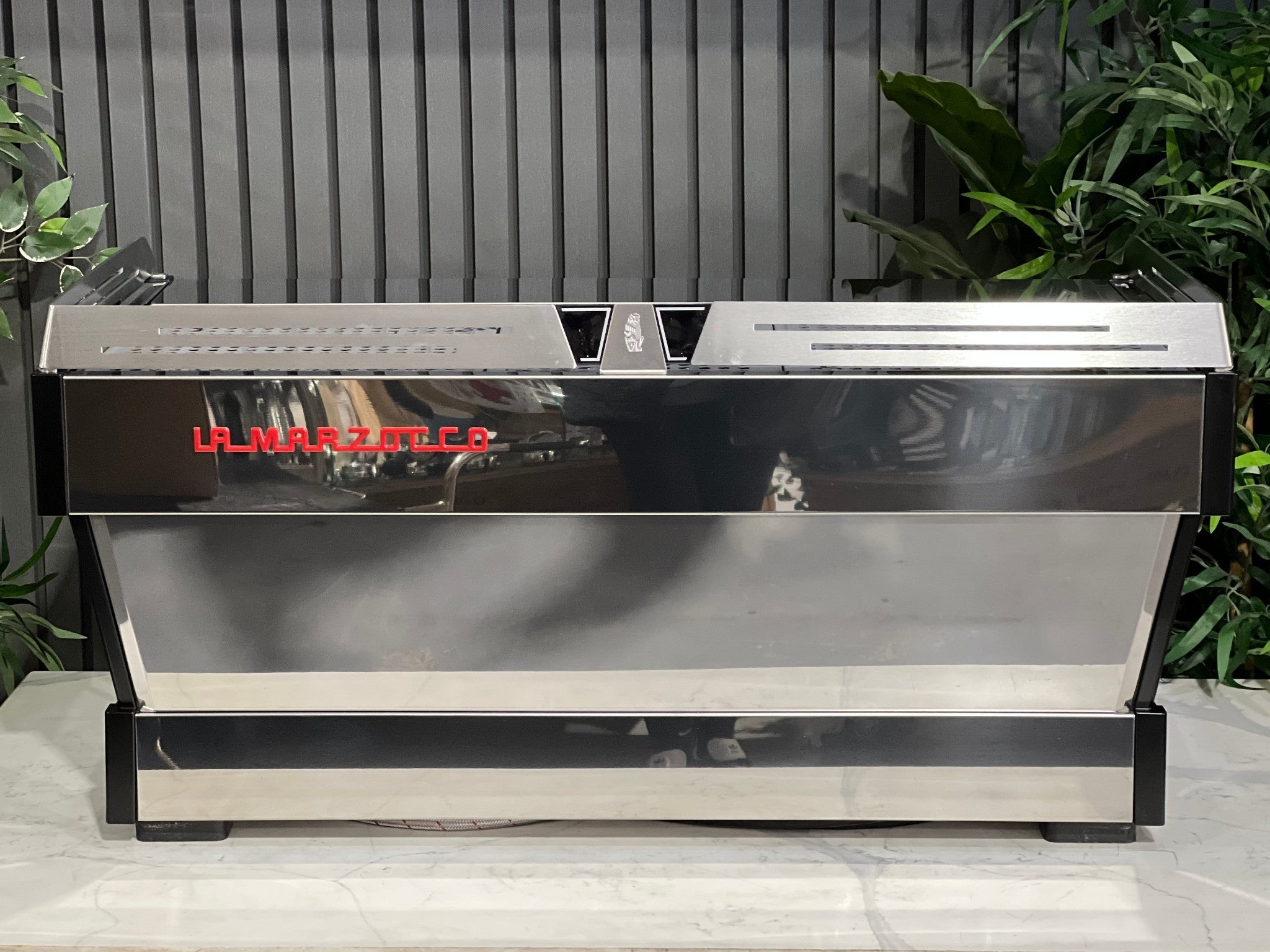 La Marzocco Linea PB ABR 3 Group Coffee Machine Stainless - New, back view showing stainless steel body and red La Marzocco logo, positioned on a marble bench with greenery in the background.