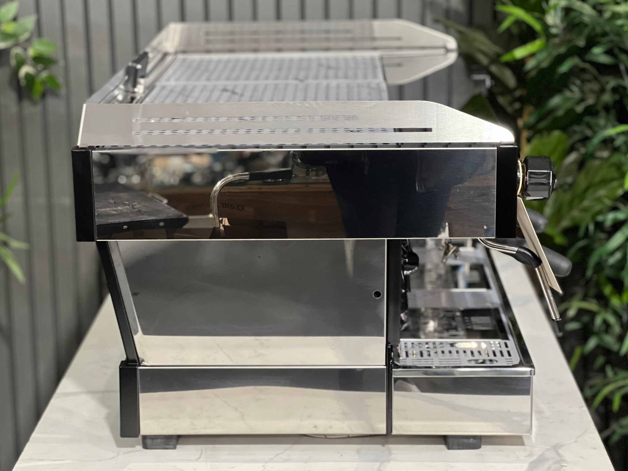 La Marzocco Linea PB ABR 3 Group Coffee Machine Stainless - New, side view showing stainless steel body, dual steam wands, and integrated ABR drip tray, set on a marble bench with greenery behind.