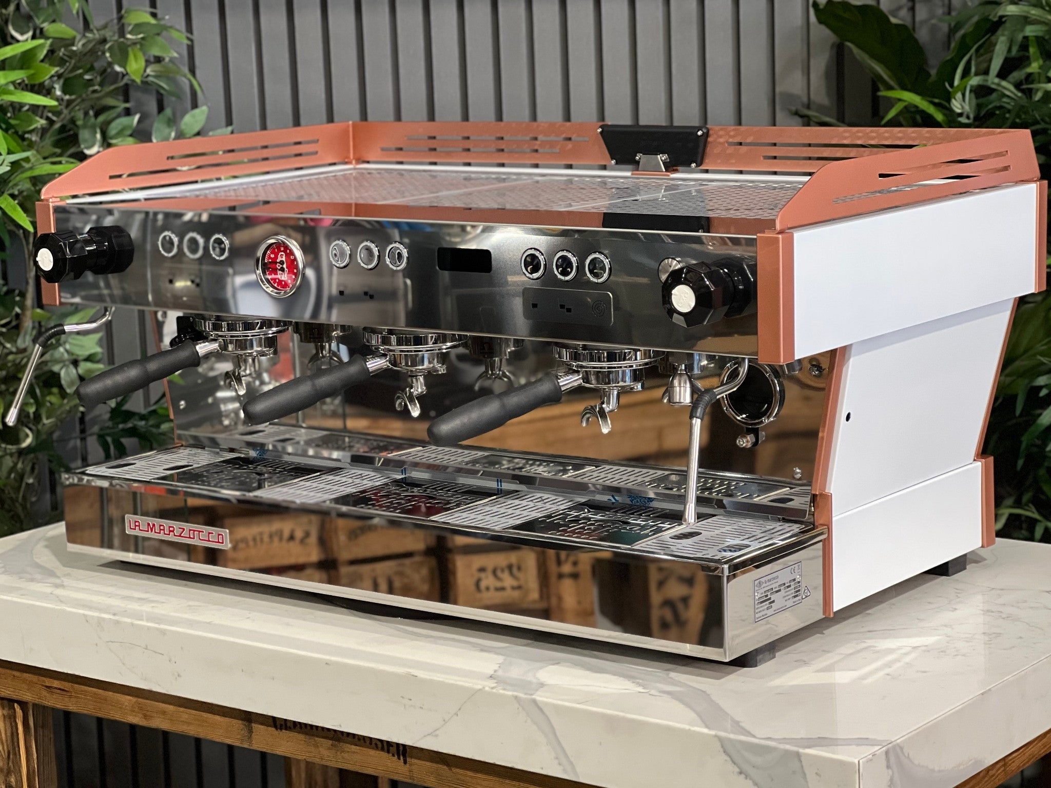 La Marzocco Linea PB ABR 3 Group Coffee Machine White & Copper - New angled view showing its white body with copper accents, dual boiler system, three saturated group heads, dual steam wands, integrated ABR drip tray, digital LED display, and barista lights on a marble bench with greenery in the background.