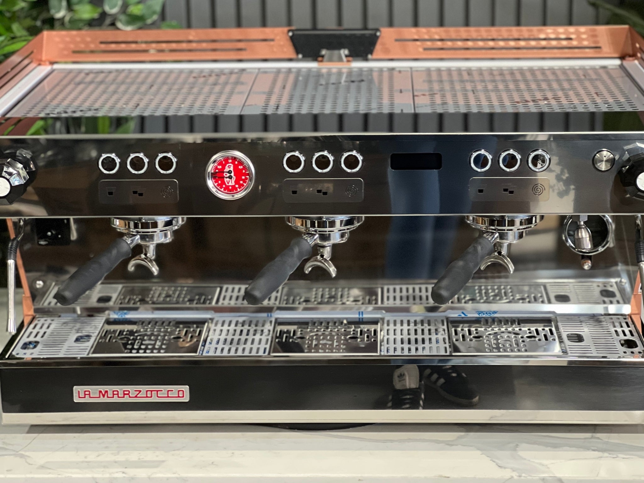 La Marzocco Linea PB ABR 3 Group Coffee Machine White & Copper - New close-up front view of the three saturated group heads resting on the ABR drip tray with integrated scales, with copper accents and part of the digital LED display visible, on a marble bench with greenery.