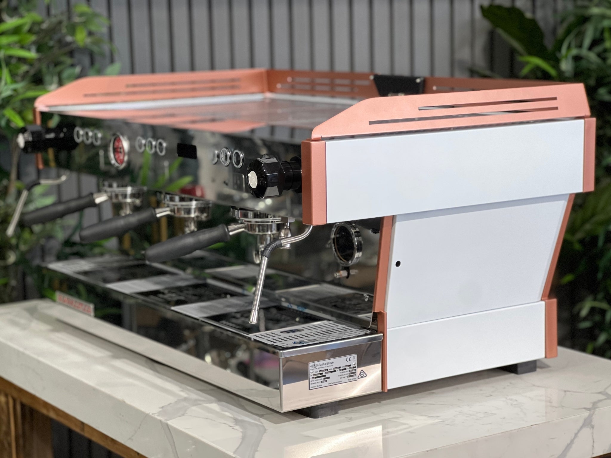 La Marzocco Linea PB ABR 3 Group Coffee Machine White & Copper - New angled view showing its white body with copper accents, dual boiler system, three saturated group heads, dual steam wands, integrated ABR drip tray, digital LED display, and barista lights on a marble bench with greenery in the background.