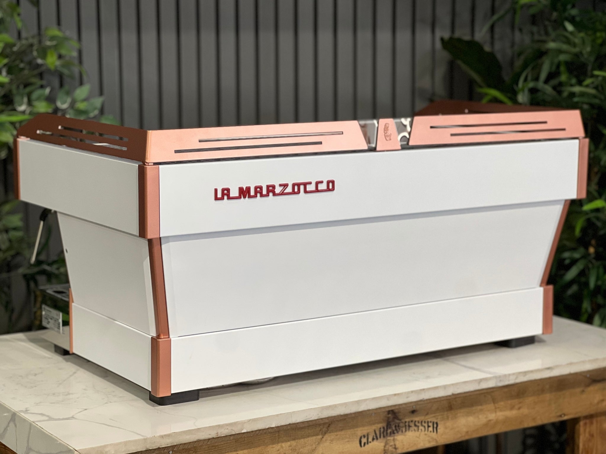 La Marzocco Linea PB ABR 3 Group Coffee Machine White & Copper - New angled back view showing piping, stainless steel rear panels, red La Marzocco logo, and dual boiler system, positioned on a marble bench with greenery behind.