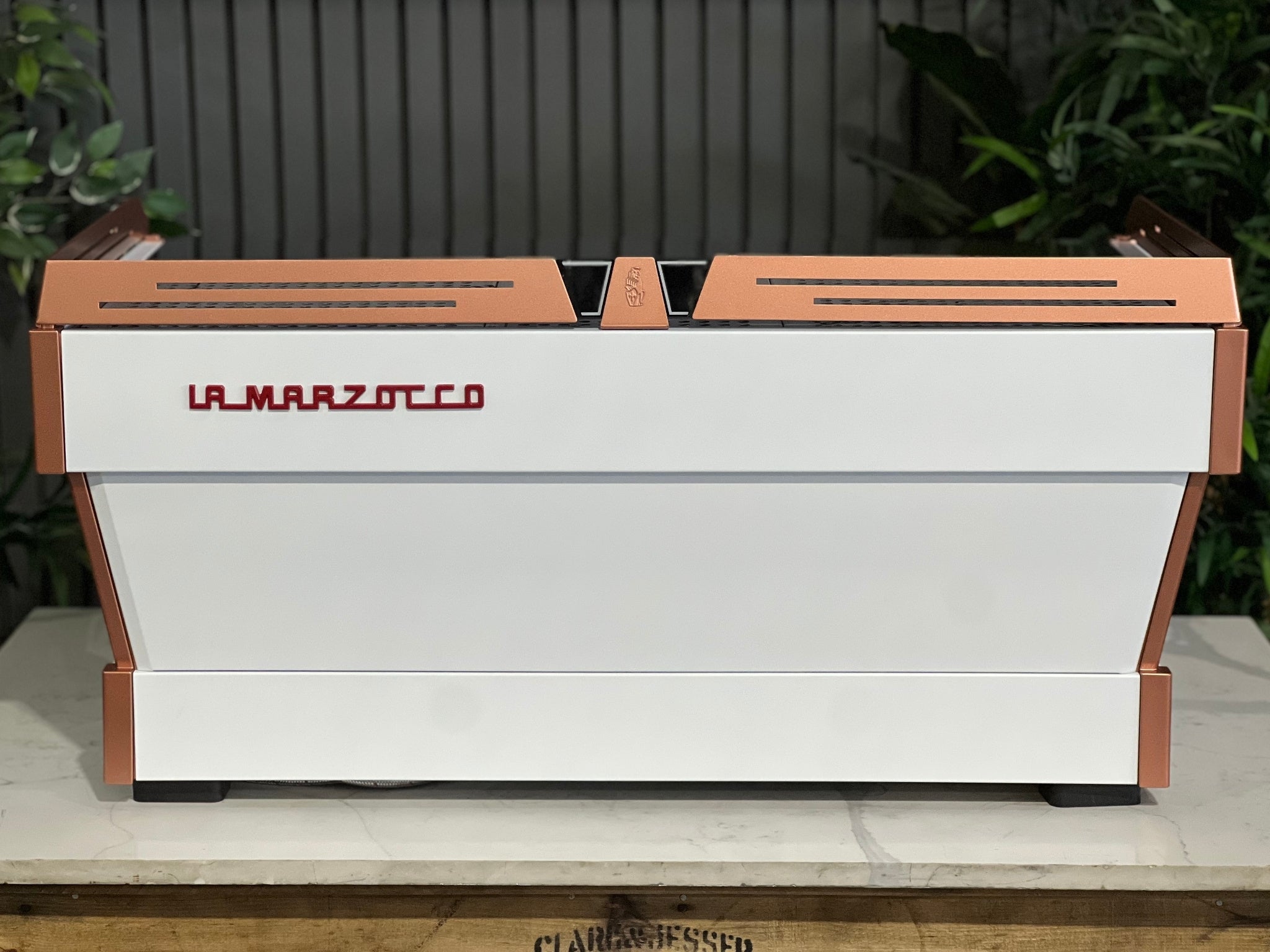 La Marzocco Linea PB ABR 3 Group Coffee Machine White & Copper - New rear view showing stainless steel panels, piping, red La Marzocco logo, and dual boiler setup, captured on a marble bench with greenery in the background.