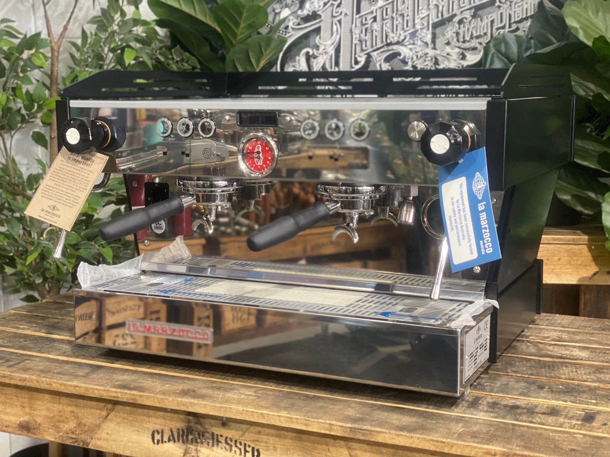 La Marzocco Linea PB 2 Group Coffee Machine Black - New angled view showing the matte black espresso machine body, dual group heads, dual stainless steel steam wands, barista lights above the groups, and cup tray on a wooden bench with greenery in the background.