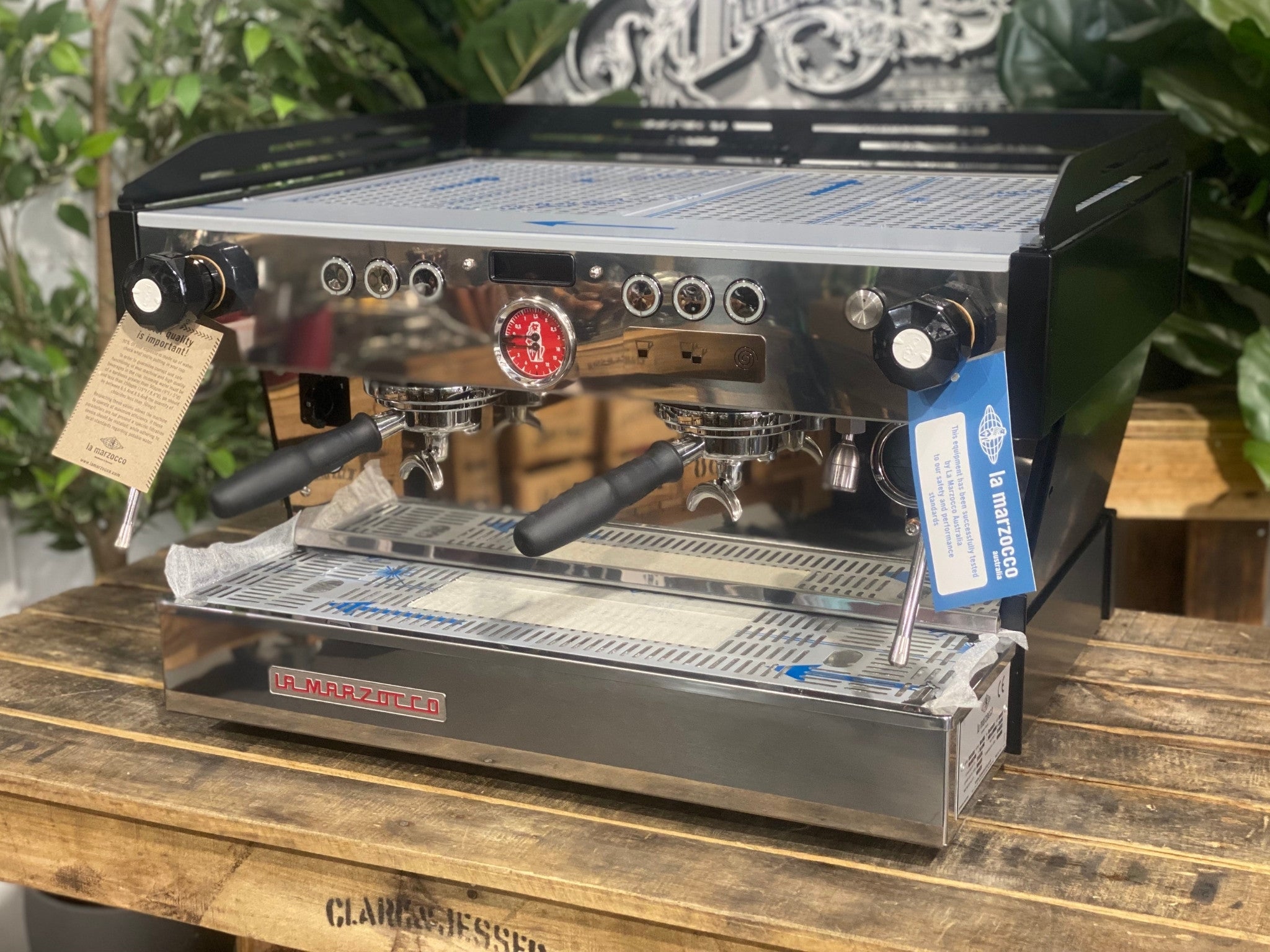 La Marzocco Linea PB 2 Group Coffee Machine Black - New angled view showing the matte black espresso machine body, dual group heads, dual stainless steel steam wands, barista lights above the groups, and cup tray on a wooden bench with greenery in the background.