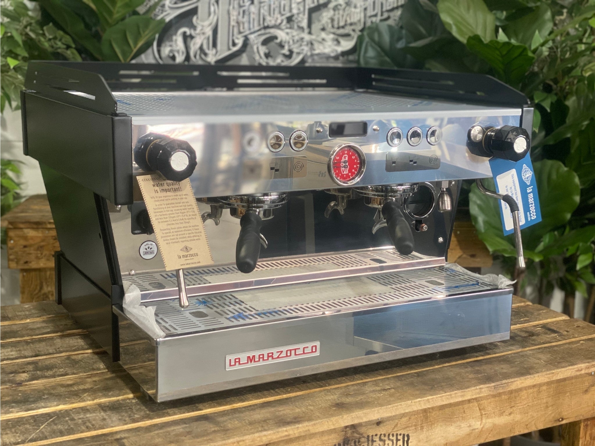 La Marzocco Linea PB 2 Group Coffee Machine Black - New angled view showing the matte black espresso machine body, dual group heads, dual stainless steel steam wands, barista lights above the groups, and cup tray on a wooden bench with greenery in the background.