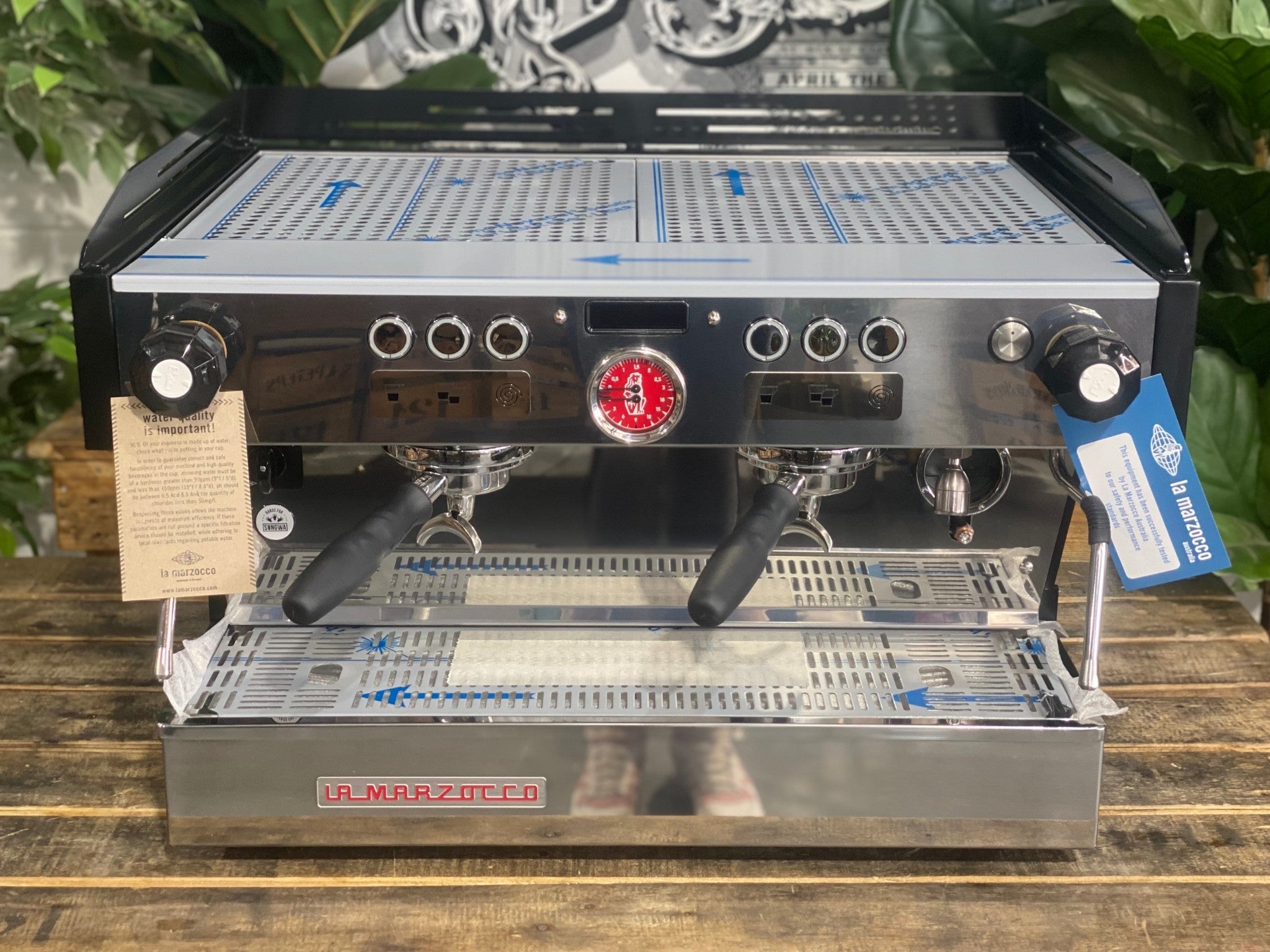 La Marzocco Linea PB 2 Group Coffee Machine Black - New front-on view from above showing the espresso machine’s black body, dual group heads, dual steam wands, barista lights, and cup tray on a wooden bench with greenery in the background.