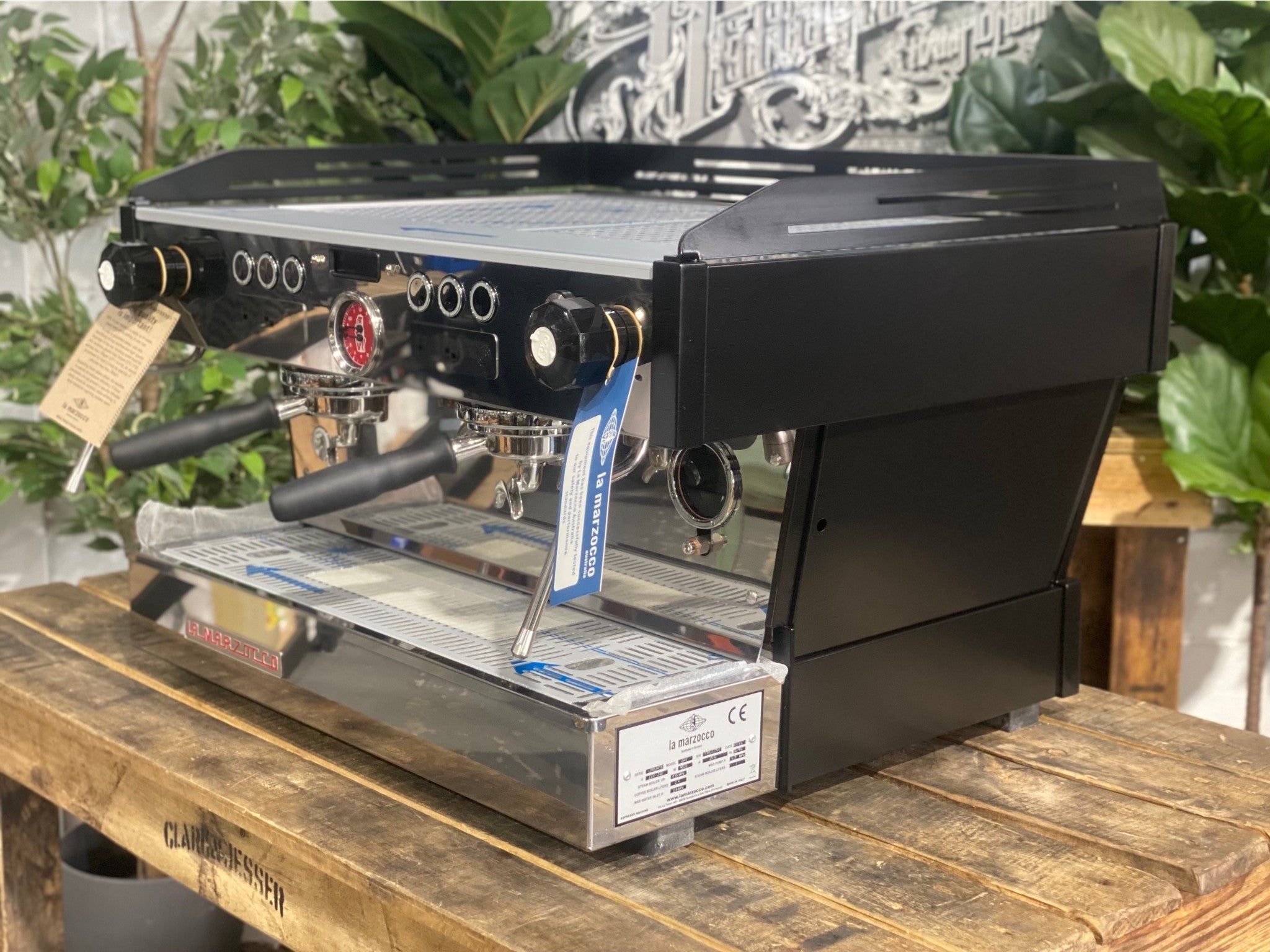 La Marzocco Linea PB 2 Group Coffee Machine Black - New angled view showing the matte black espresso machine body, dual group heads, dual stainless steel steam wands, barista lights above the groups, and cup tray on a wooden bench with greenery in the background.