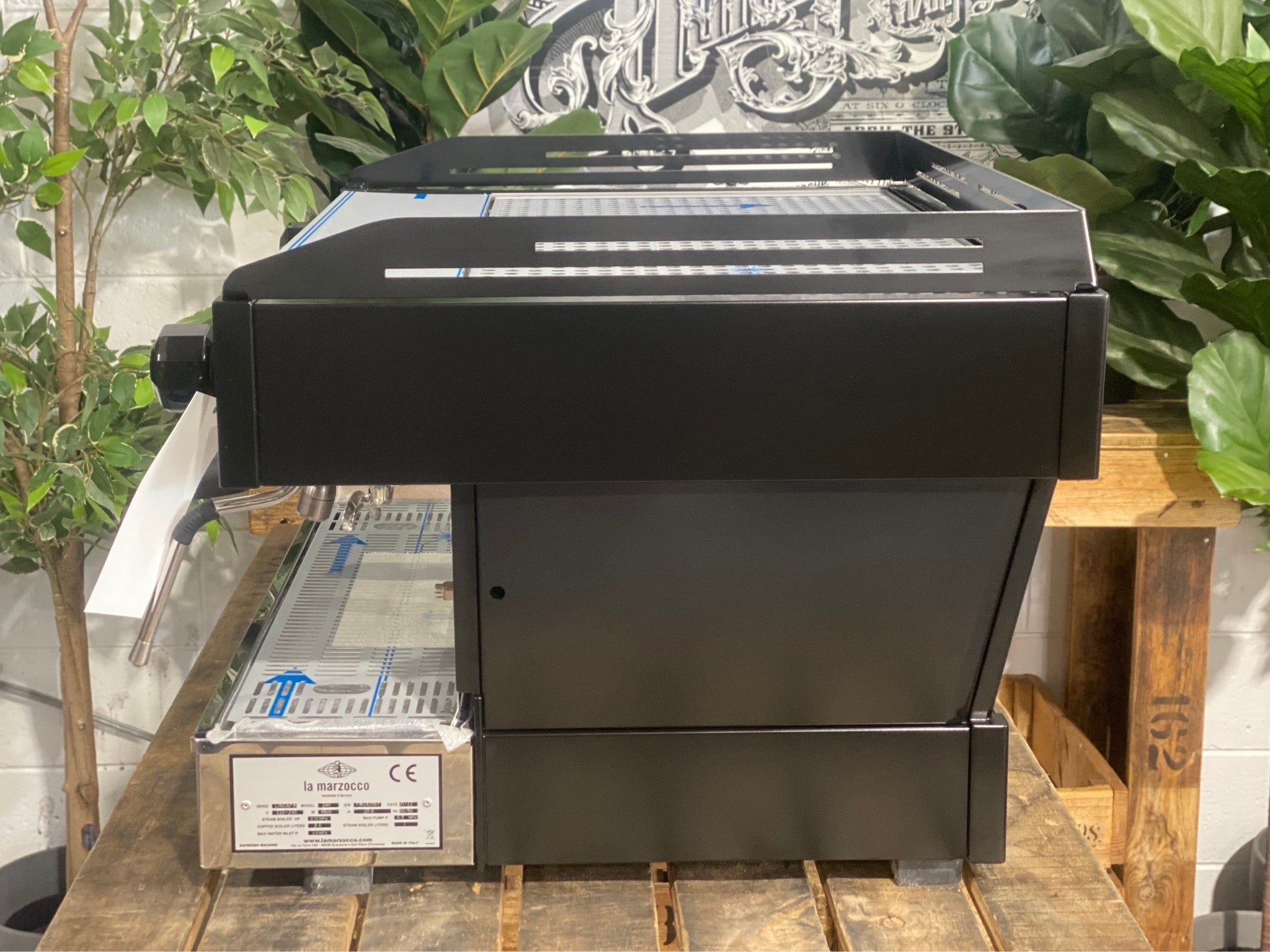 La Marzocco Linea PB 2 Group Coffee Machine Black - New side view showing the sleek black espresso machine body, side panels, and steam wand base, on a wooden bench with greenery in the background.