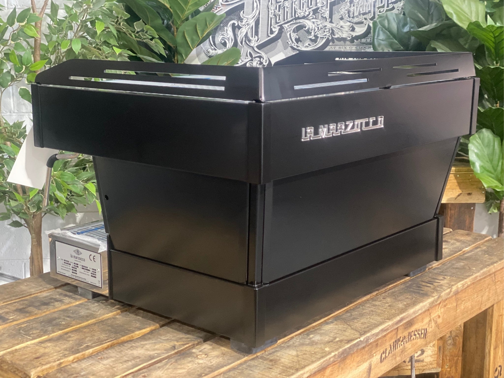 La Marzocco Linea PB 2 Group Coffee Machine Black - New angled back view highlighting the black espresso machine’s body panels and overall shape on a wooden bench with greenery in the background.