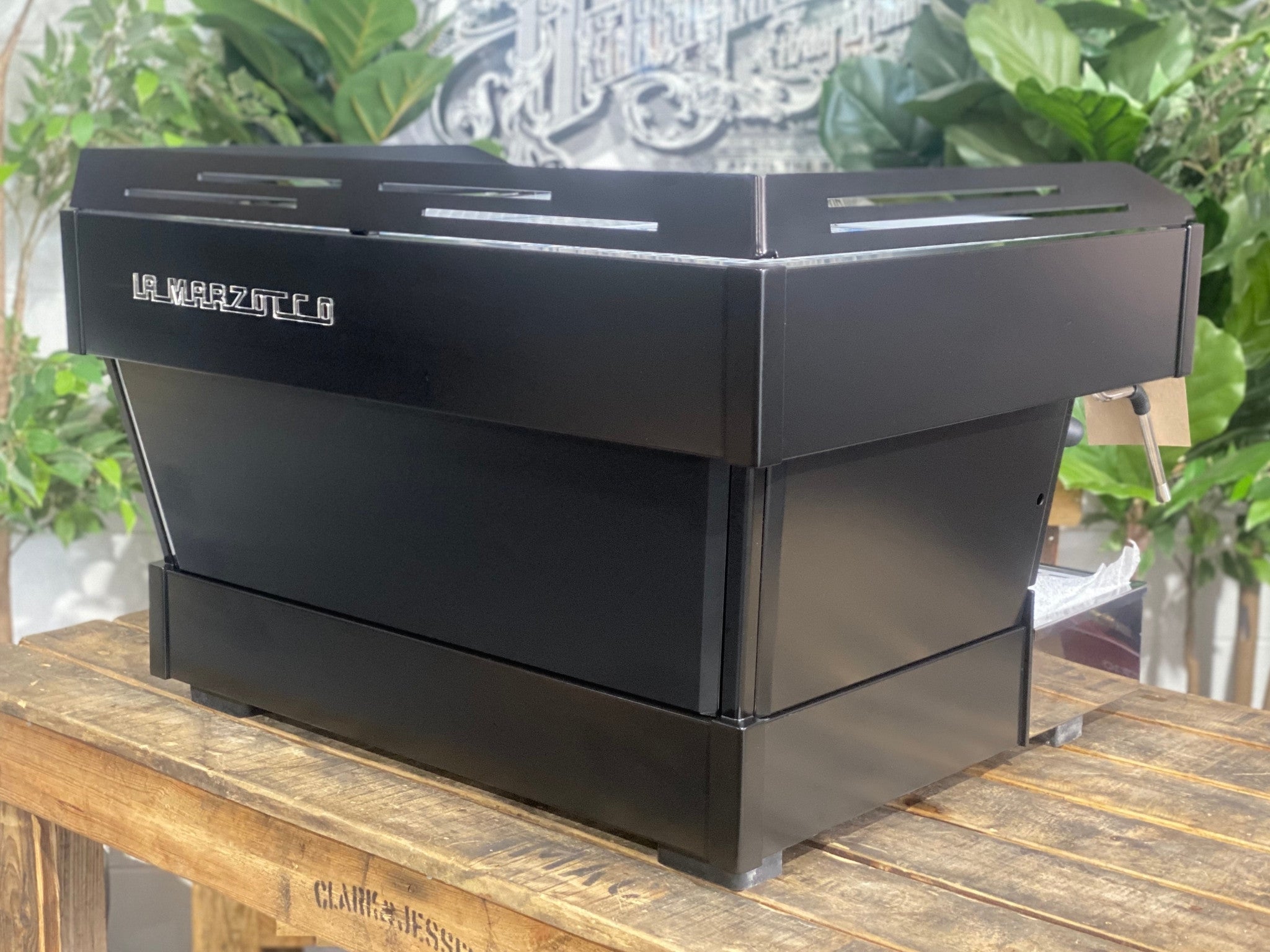 La Marzocco Linea PB 2 Group Coffee Machine Black - New angled back view highlighting the black espresso machine’s body panels and overall shape on a wooden bench with greenery in the background.