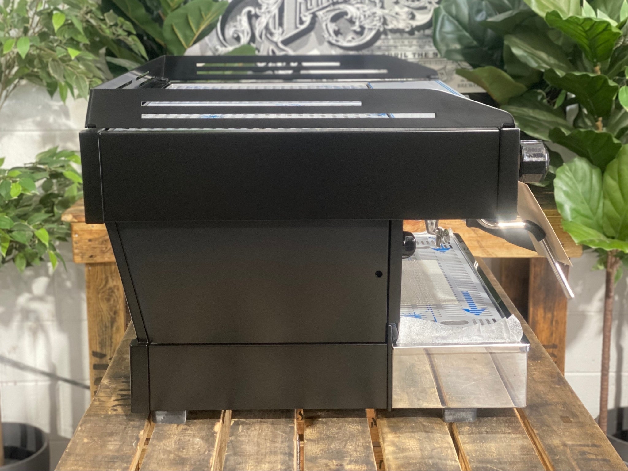 La Marzocco Linea PB 2 Group Coffee Machine Black - New side view showing the sleek black espresso machine body, side panels, and steam wand base, on a wooden bench with greenery in the background.