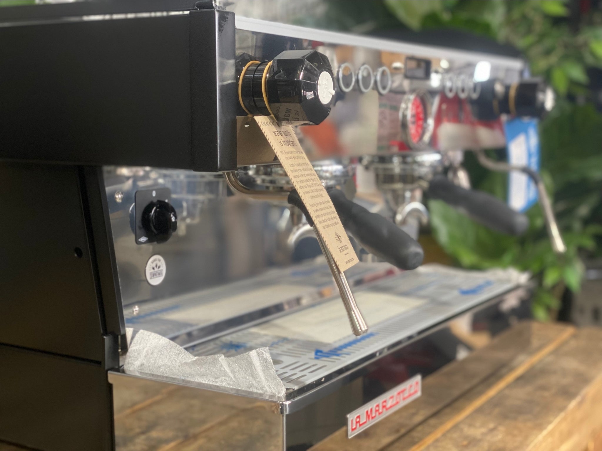 La Marzocco Linea PB 2 Group Coffee Machine Black - New close-up front view showing the espresso machine’s dual group heads, dual steam wands, barista lights, and cup tray, highlighting the black body on a wooden bench with greenery in the background.