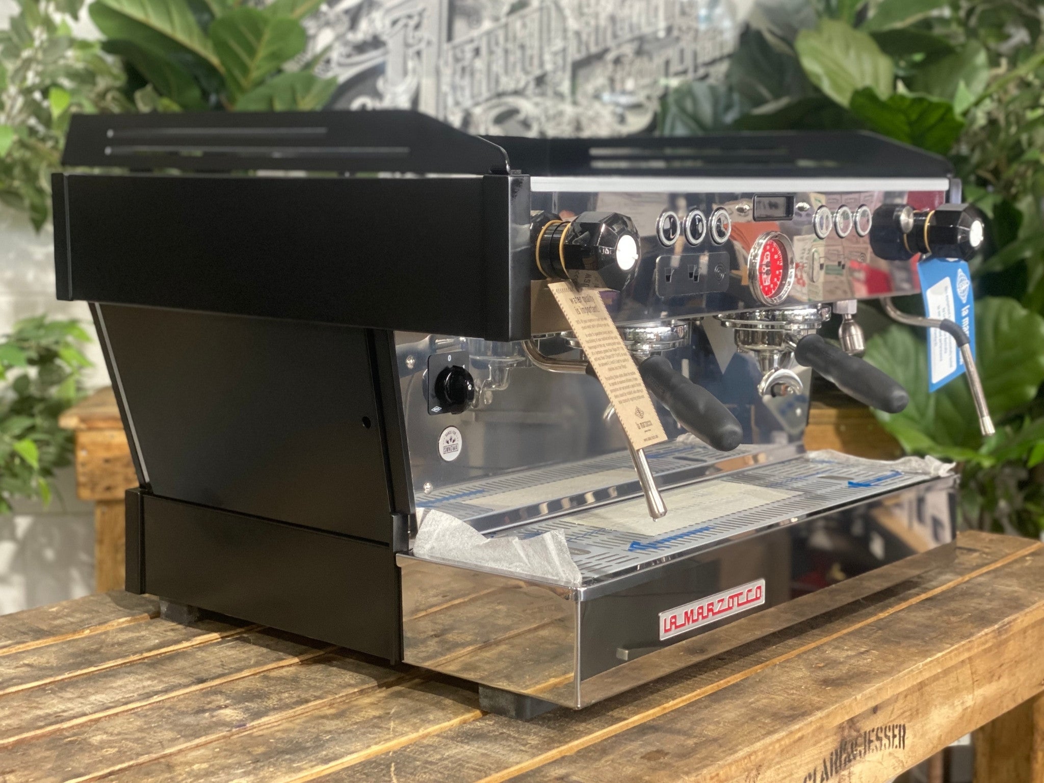 La Marzocco Linea PB 2 Group Coffee Machine Black - New angled view showing the matte black espresso machine body, dual group heads, dual stainless steel steam wands, barista lights above the groups, and cup tray on a wooden bench with greenery in the background.