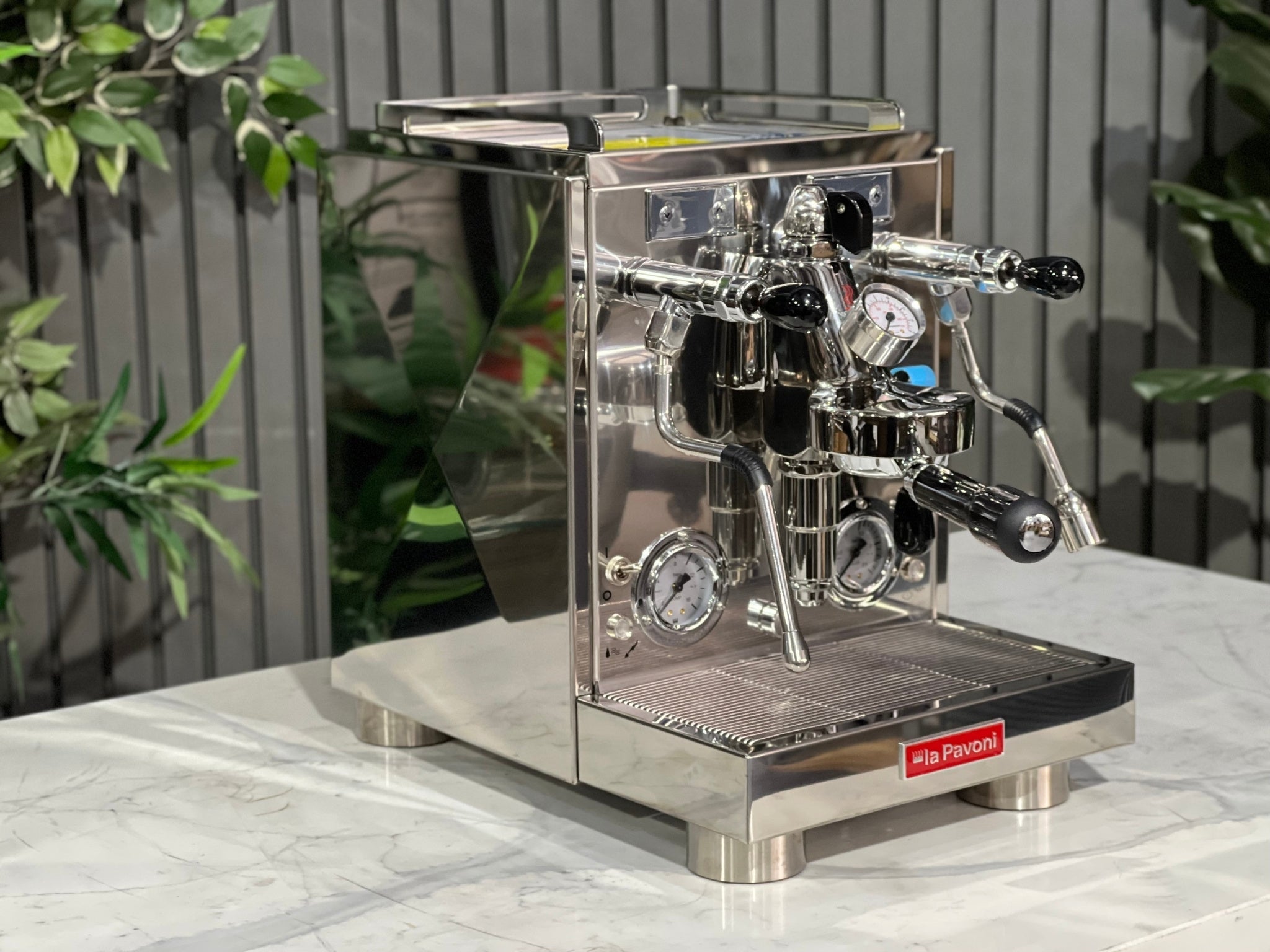 A new La Pavoni Botticelli Specialty Dual Boiler PID 1 Group Espresso Coffee Machine is sitting on a marble bench. There is greenery in the background. The image is taken from the front of the machine and the machine is sitting on an angle so that you can see the front of the machine and the side.