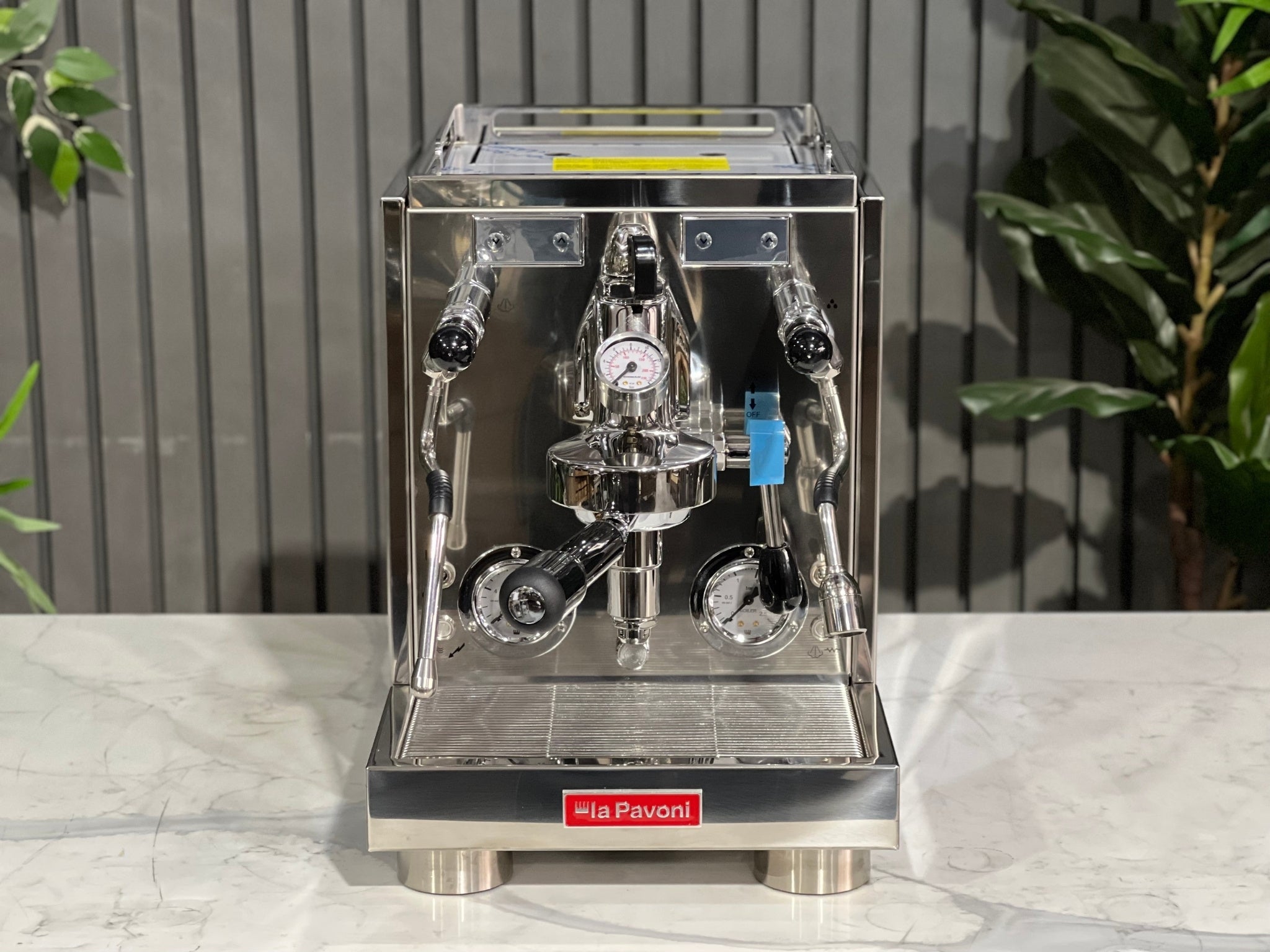A new La Pavoni Botticelli Specialty Dual Boiler PID 1 Group Espresso Coffee Machine sitting on a marble bench. There is greenery in the background. The image is taken from the front of the machine so that you can see the front of the machine, control panel, buttons and group heads.