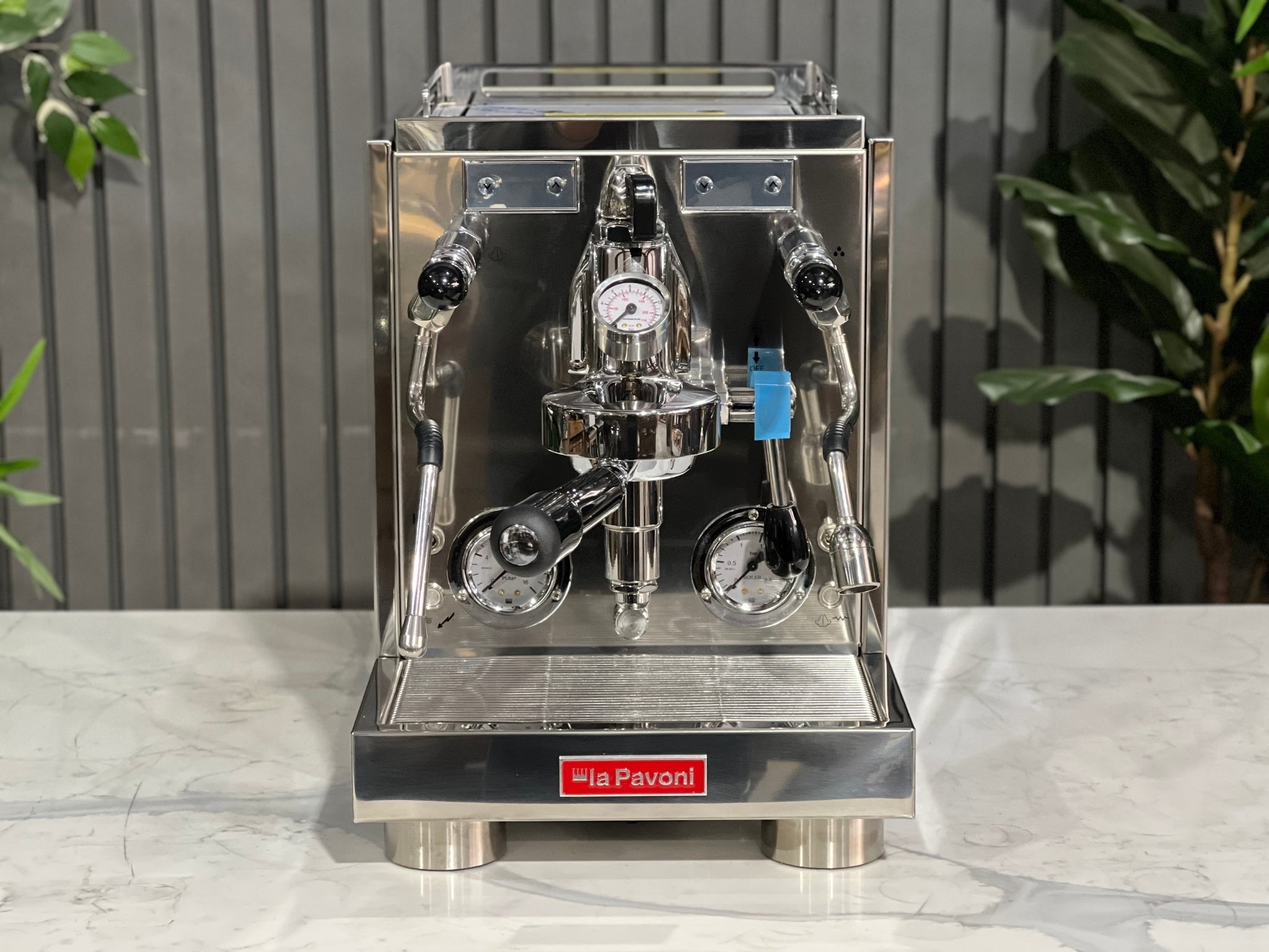A new La Pavoni Botticelli Specialty Dual Boiler PID 1 Group Espresso Coffee Machine sitting on a marble bench. There is greenery in the background. The image is taken from the front of the machine so that you can see the front of the machine, controls, buttons and group head.