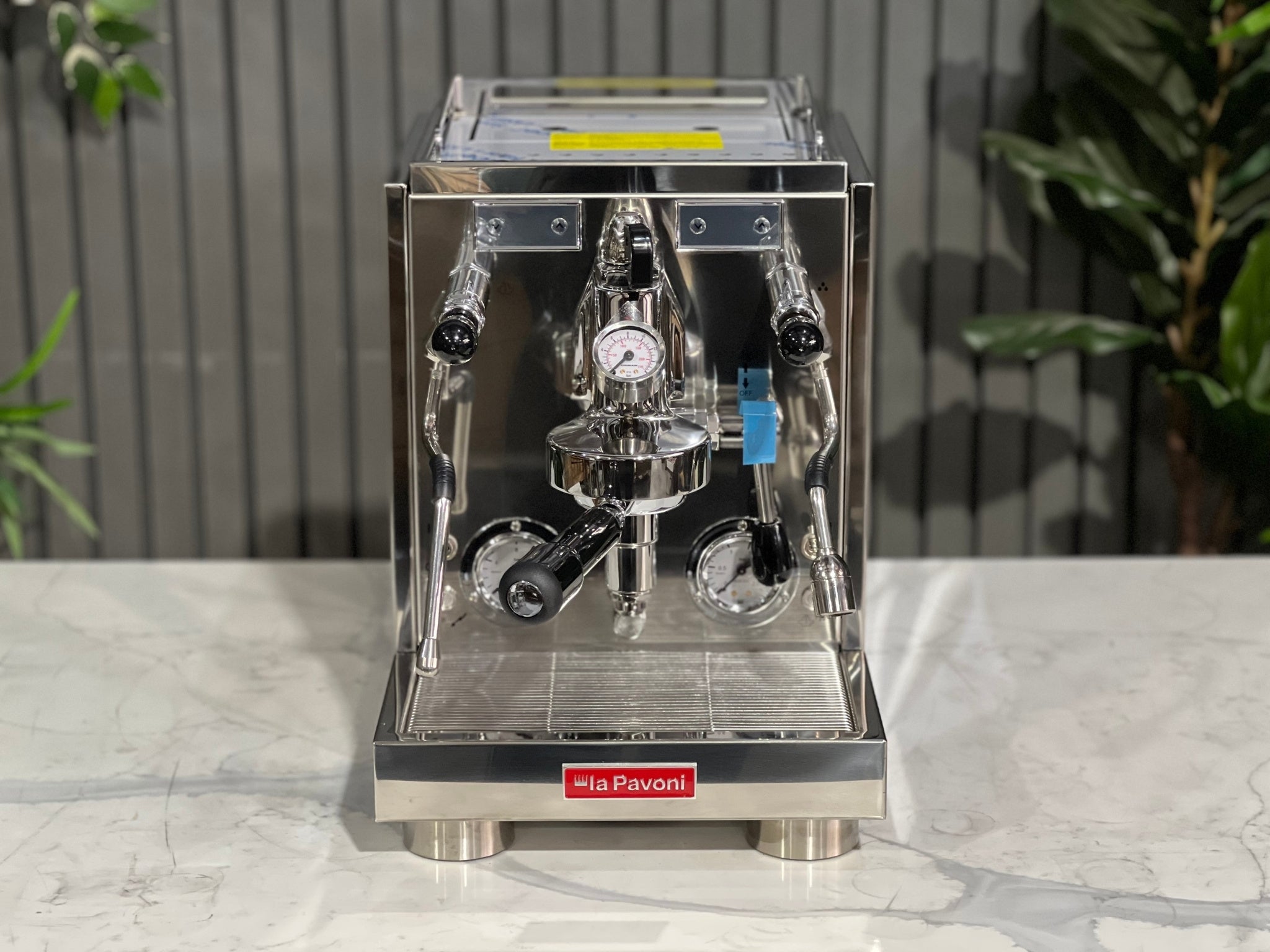 A new La Pavoni Botticelli Specialty Dual Boiler PID 1 Group Espresso Coffee Machine sitting on a marble bench. There is greenery in the background. The image is taken from the front of the machine so that you can see the front of the machine, control panel, buttons and group heads.