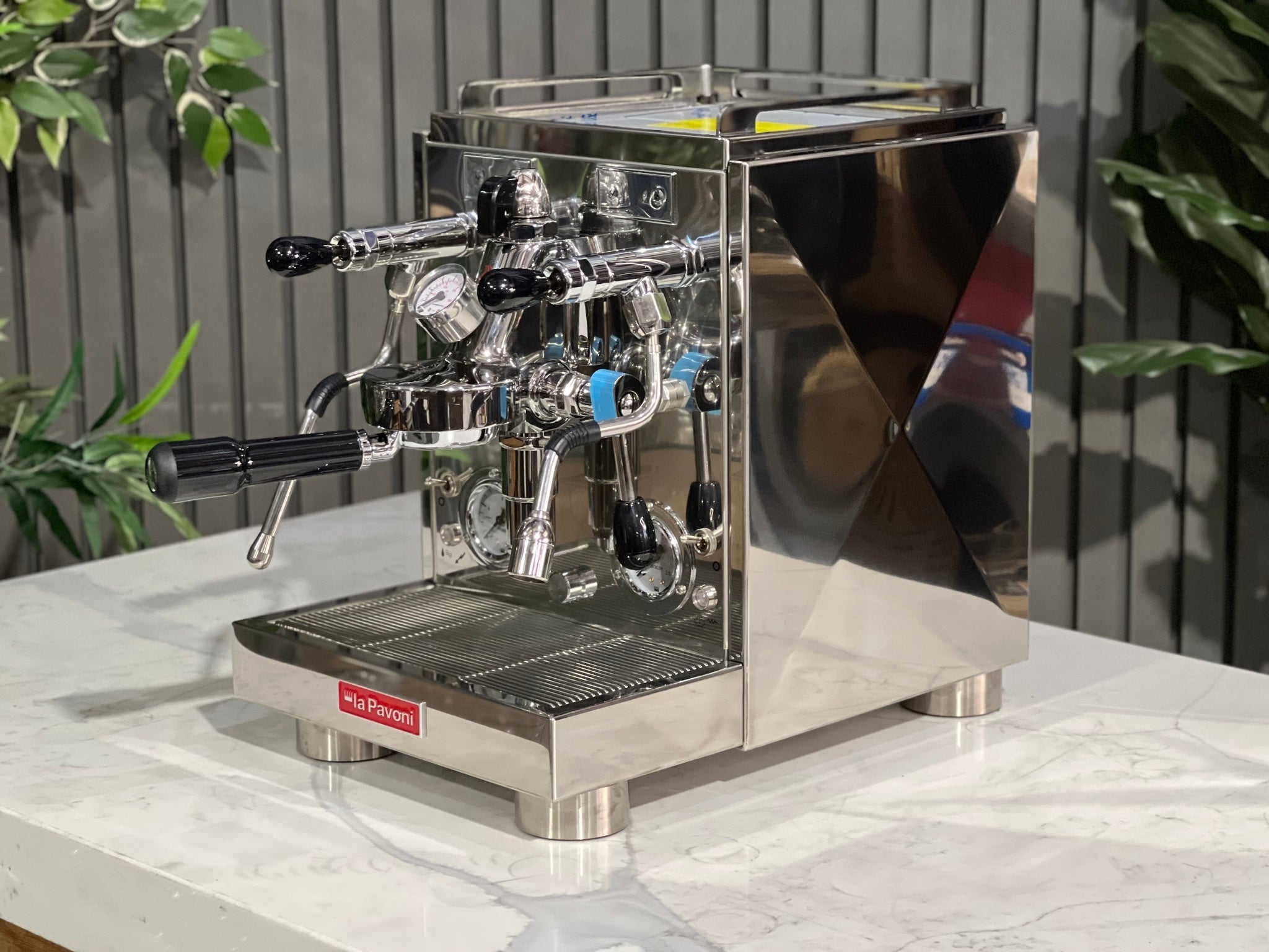 A new La Pavoni Botticelli Specialty Dual Boiler PID 1 Group Espresso Coffee Machine is sitting on a marble bench. There is greenery in the background. The image is taken from the front of the machine and the machine is sitting on an angle so that you can see the front of the machine and the side.