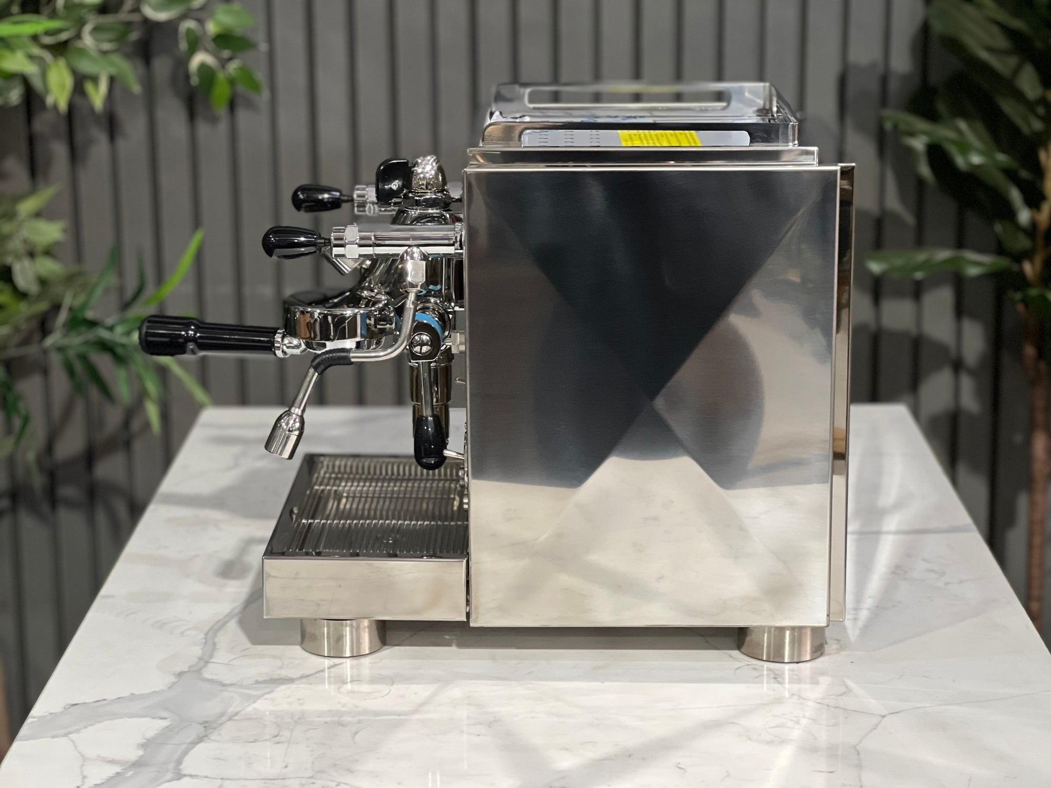 A new La Pavoni Botticelli Specialty Dual Boiler PID 1 Group Espresso Coffee Machine sitting on a marble bench. There is greenery in the background. The image is taken from the side of the machine.