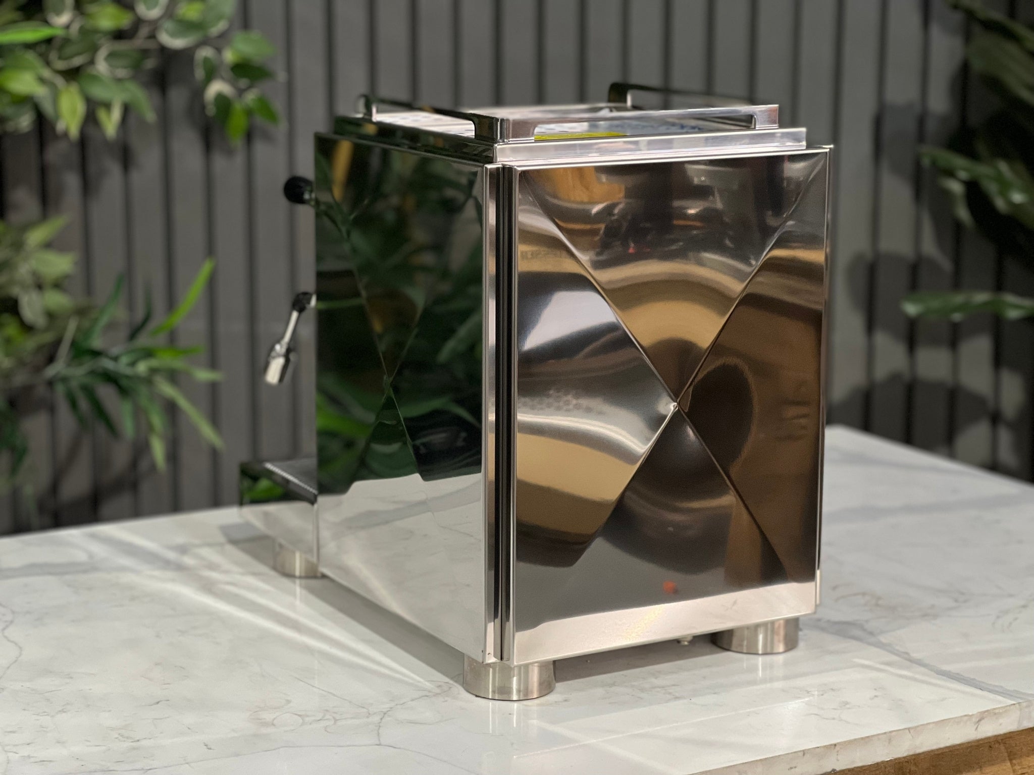 A new La Pavoni Botticelli Specialty Dual Boiler PID 1 Group Espresso Coffee Machine  sitting on a marble bench. There is greenery in the background. The image is taken from behind the machine and the machine is sitting on an angle so that you can see the back of the machine and the side.