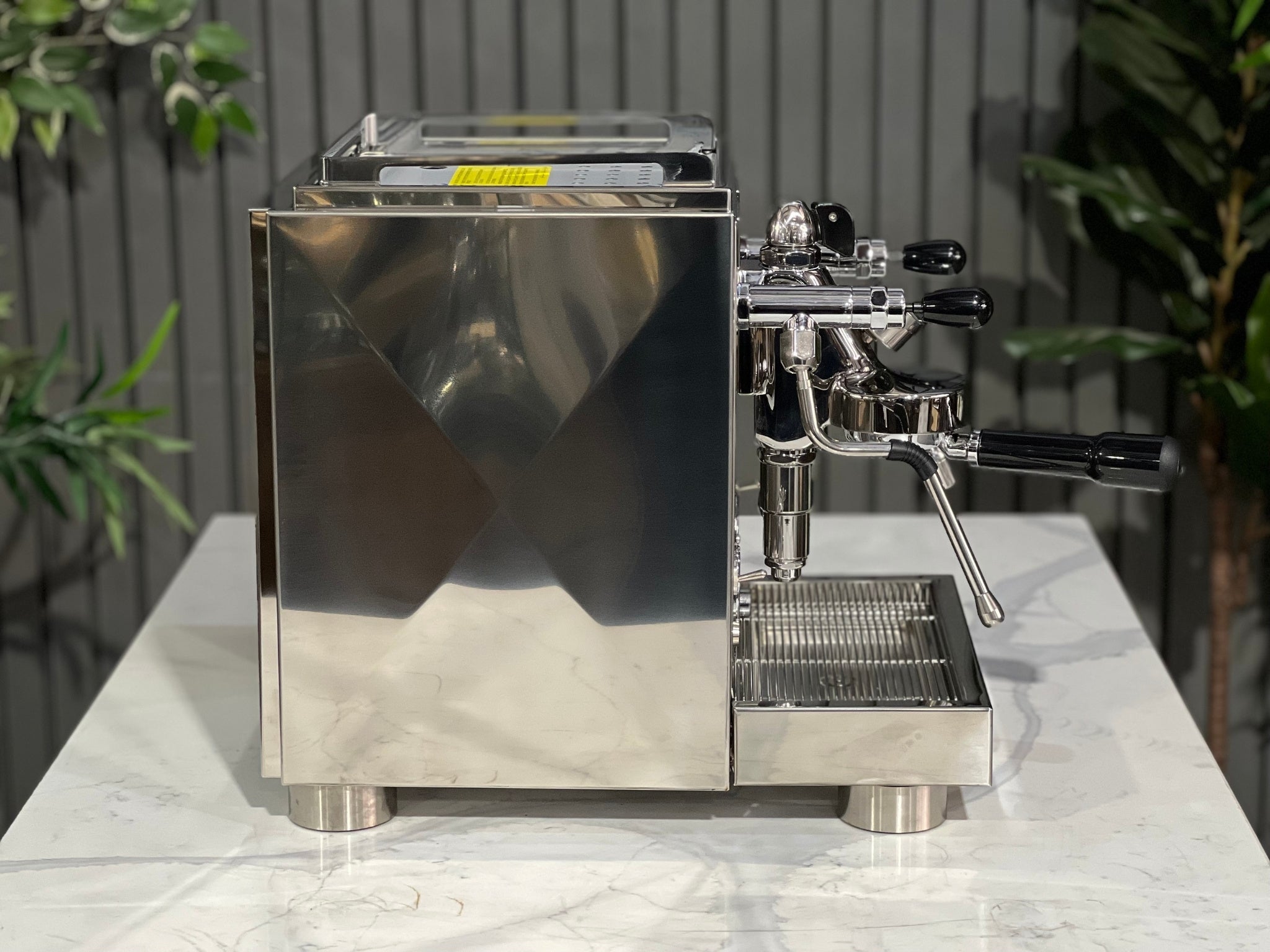 A new La Pavoni Botticelli Specialty Dual Boiler PID 1 Group Espresso Coffee Machine sitting on a marble bench. There is greenery in the background. The image is taken from the side of the machine.