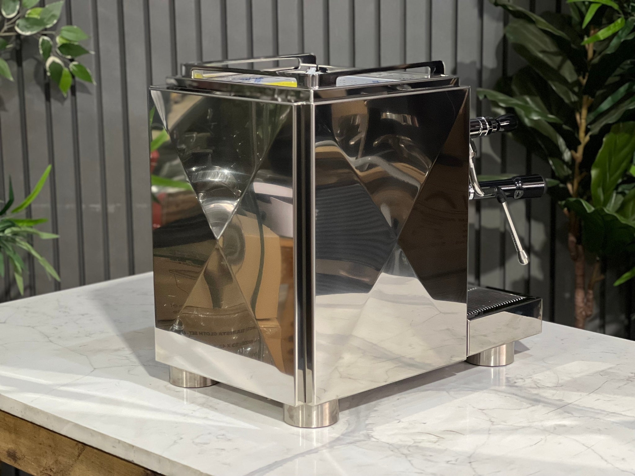 A new La Pavoni Botticelli Specialty Dual Boiler PID 1 Group Espresso Coffee Machine  sitting on a marble bench. There is greenery in the background. The image is taken from behind the machine and the machine is sitting on an angle so that you can see the back of the machine and the side.